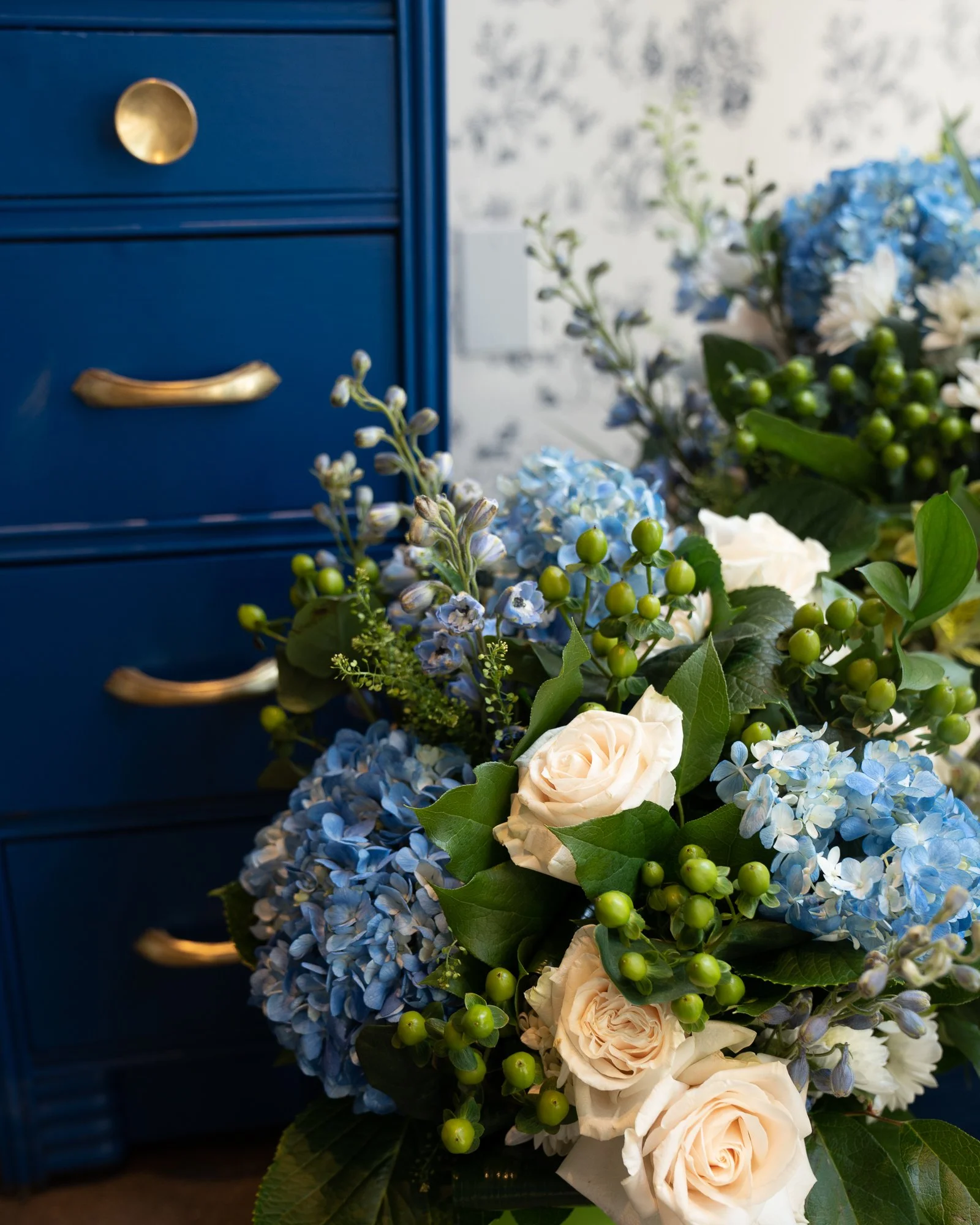 A floral arrangement with white roses, blue hydrangeas, green berries, and assorted green leaves next to a blue dresser with gold handles.