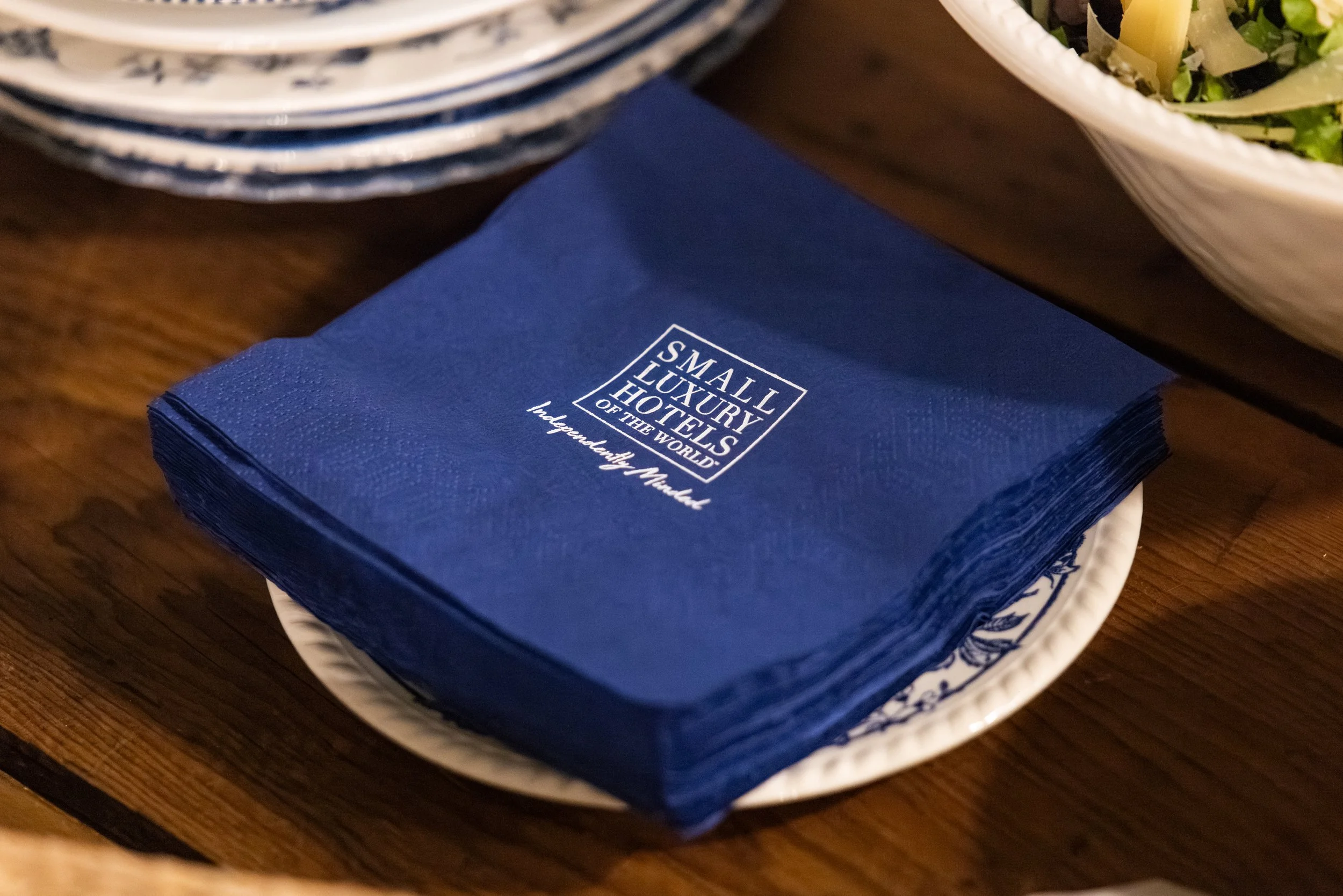 A stack of blue napkins on a white plate with blue decorative pattern, with text 'Small Luxury Hotels of the World' and 'Independency Matters' printed on them.