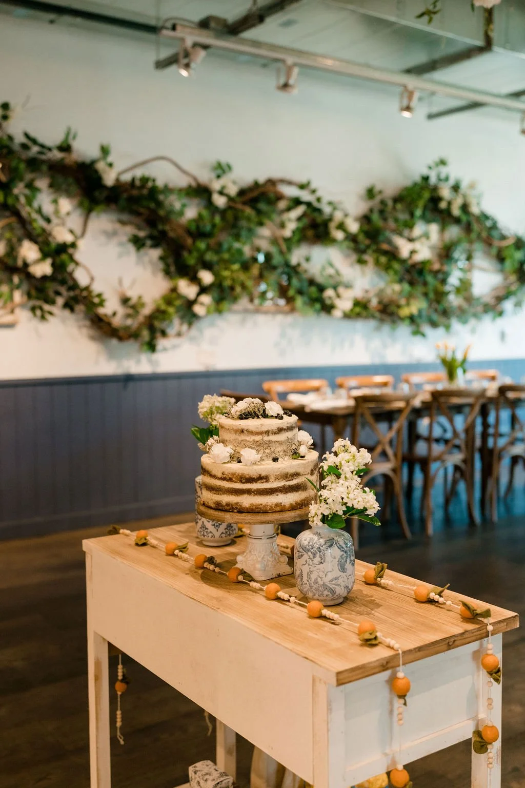 A rustic wedding cake with a semi-naked frosting style, decorated with white flowers and greenery, on a cake stand. The cake is on a light wooden table with a white base, also decorated with a string of orange and yellow beads. In the background, there are wooden chairs and a wall adorned with a large, twisted greenery and white flower arrangement.