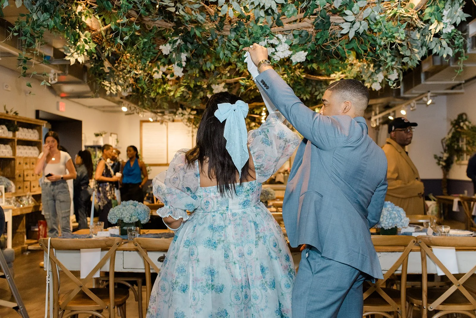 A couple dancing at an event with a decorated ceiling of greenery and white flowers, surrounded by guests in an indoor setting.