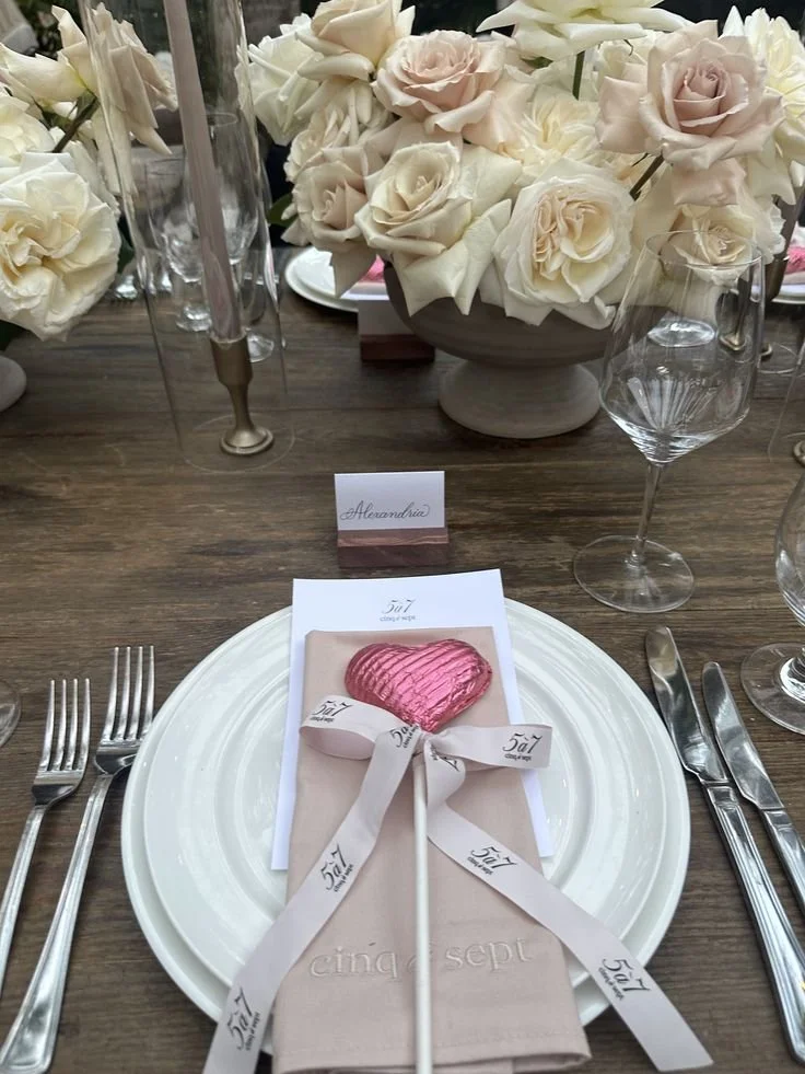 Table setting with a white plate, a pink heart-shaped lollipop, a pink napkin, and a place card labeled 'Alexandria'. Background features a large bouquet of white and cream roses, and glassware including wine glasses.