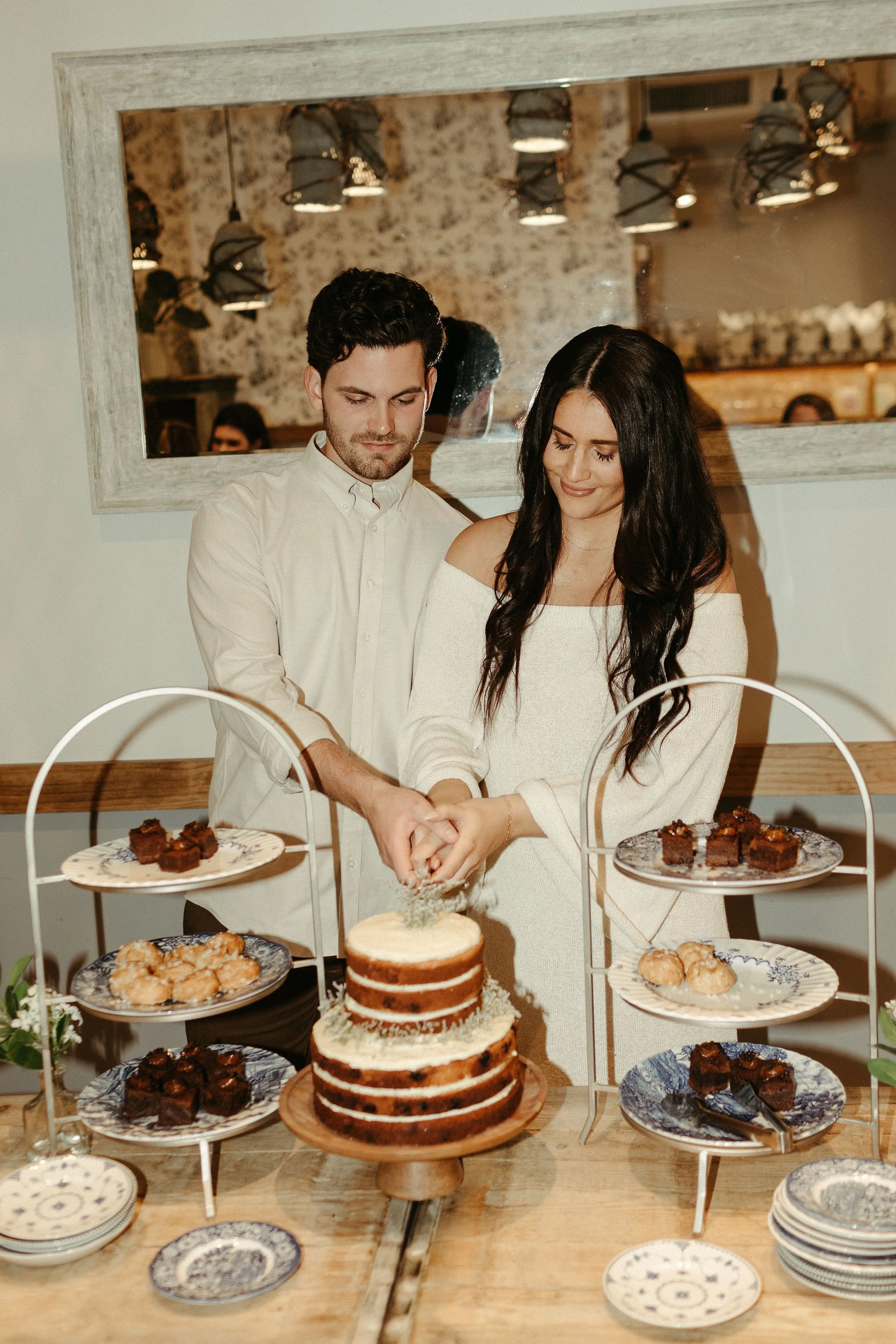 A man and woman cutting a cake at a celebration with dessert plates and baked goods on a wooden table.