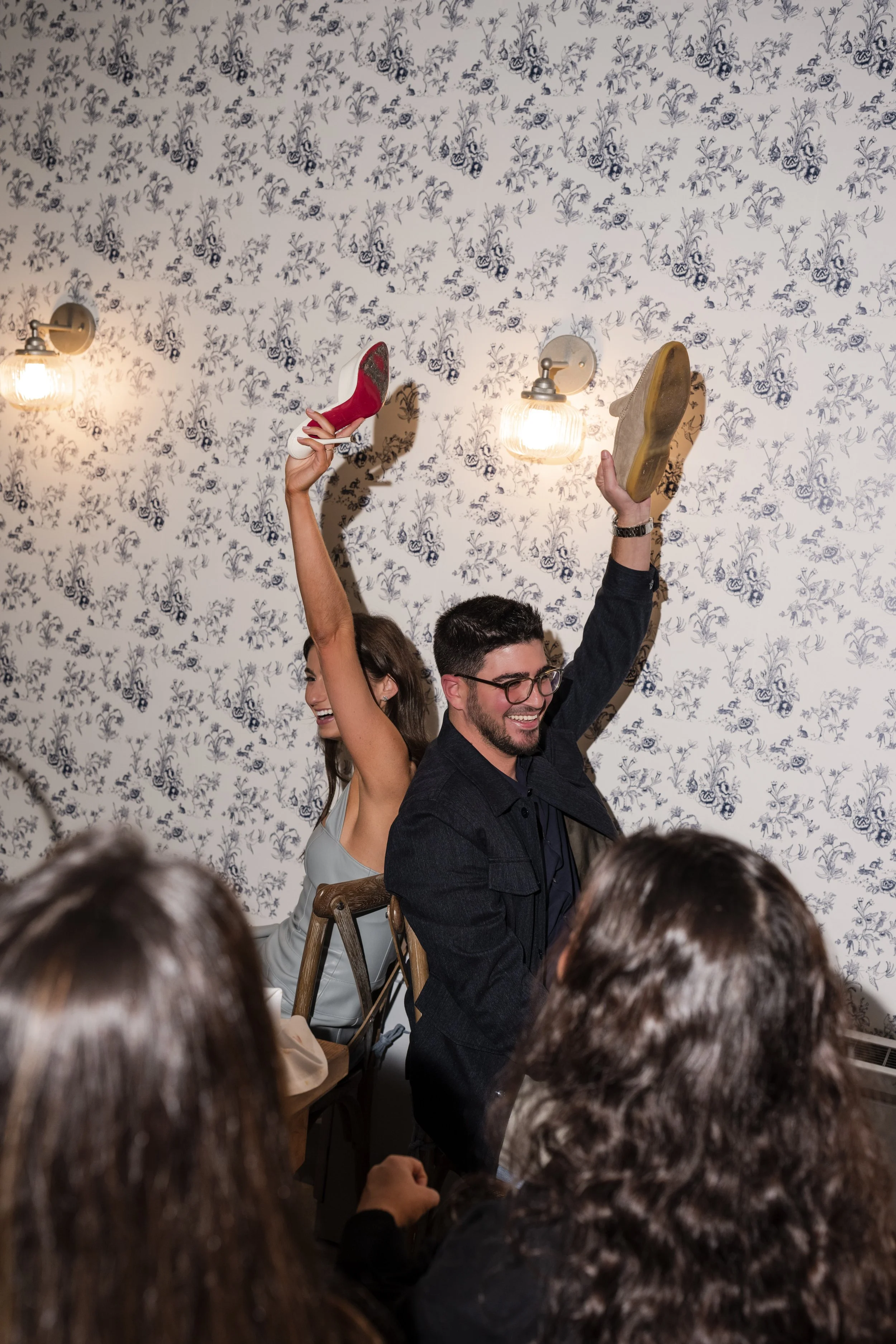 Two people celebrating at a party, one woman is raising her shoe and a plate, the man is smiling and wearing glasses, with others seen in the foreground, all smiling or laughing in a decorated room with floral wallpaper and wall-mounted lights.