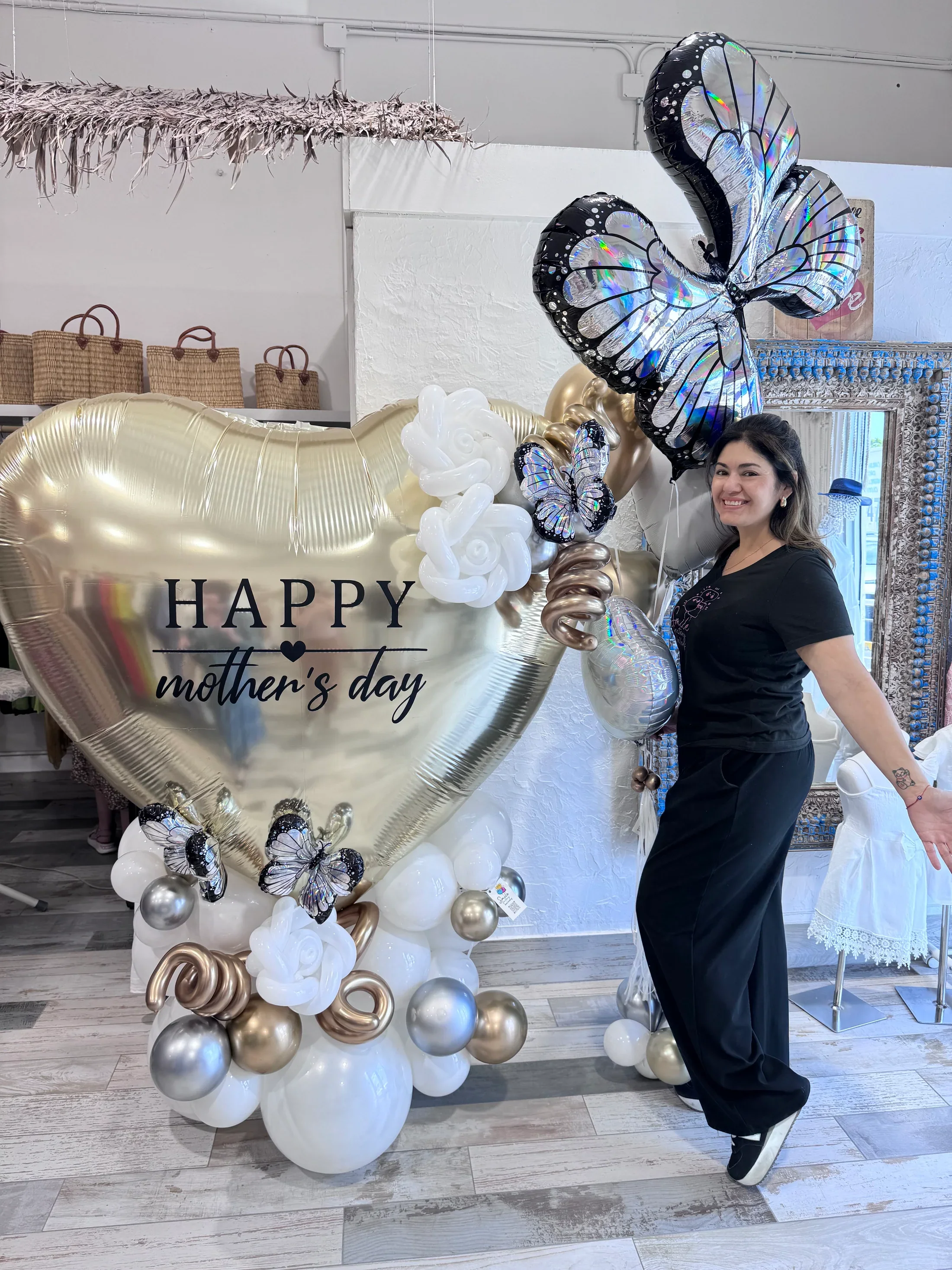 A woman standing next to a large balloon display with a gold heart-shaped balloon that says 'Happy Mother's Day,' surrounded by smaller balloons, including butterfly-shaped balloons, roses, and metallic orbs, celebrating Mother's Day.