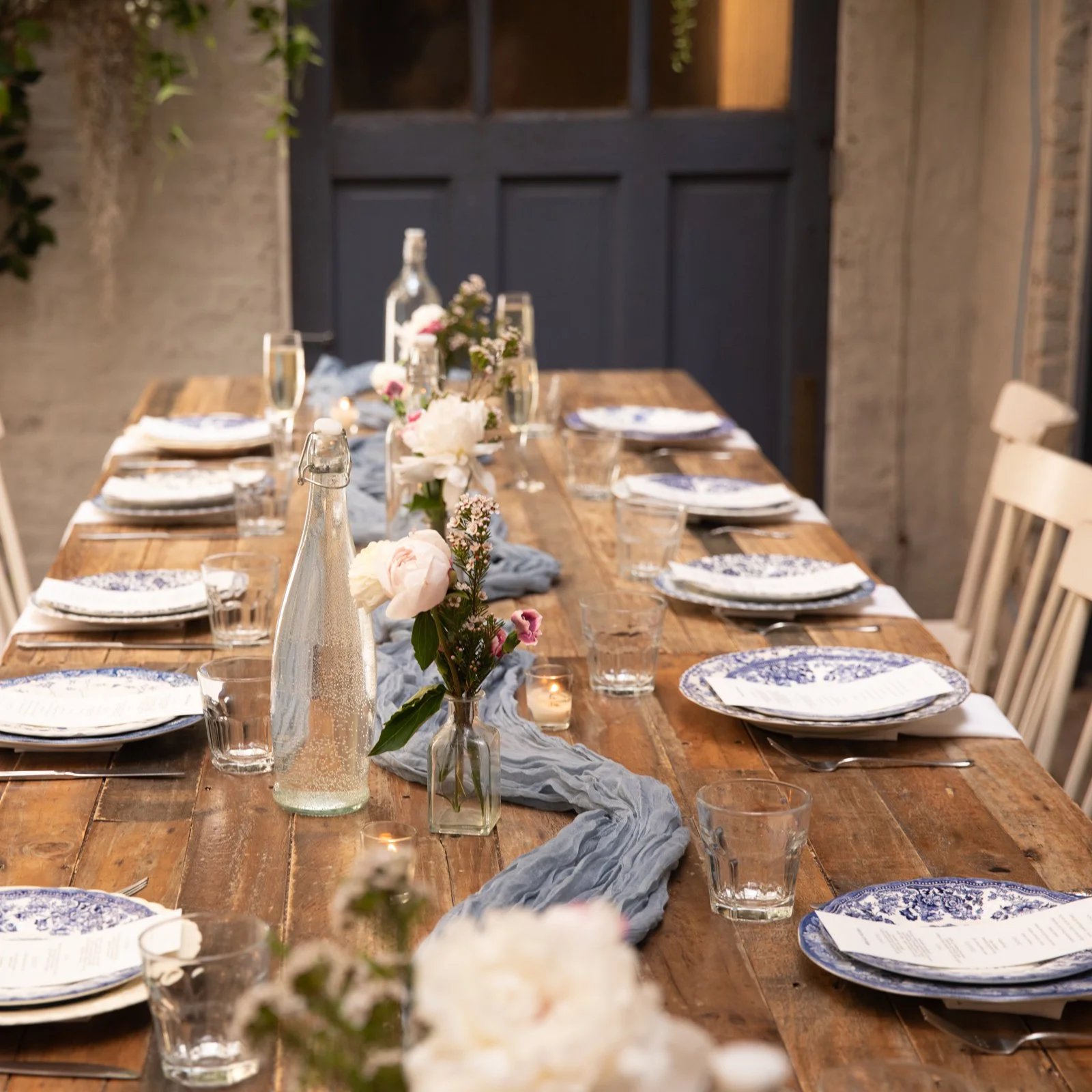 A long wooden dining table set for a meal with white and blue patterned plates, clear glasses, silverware, vases with pink and white flowers, a blue fabric table runner, and small candles in glass holders, in a rustic indoor setting with a dark door and brick walls.