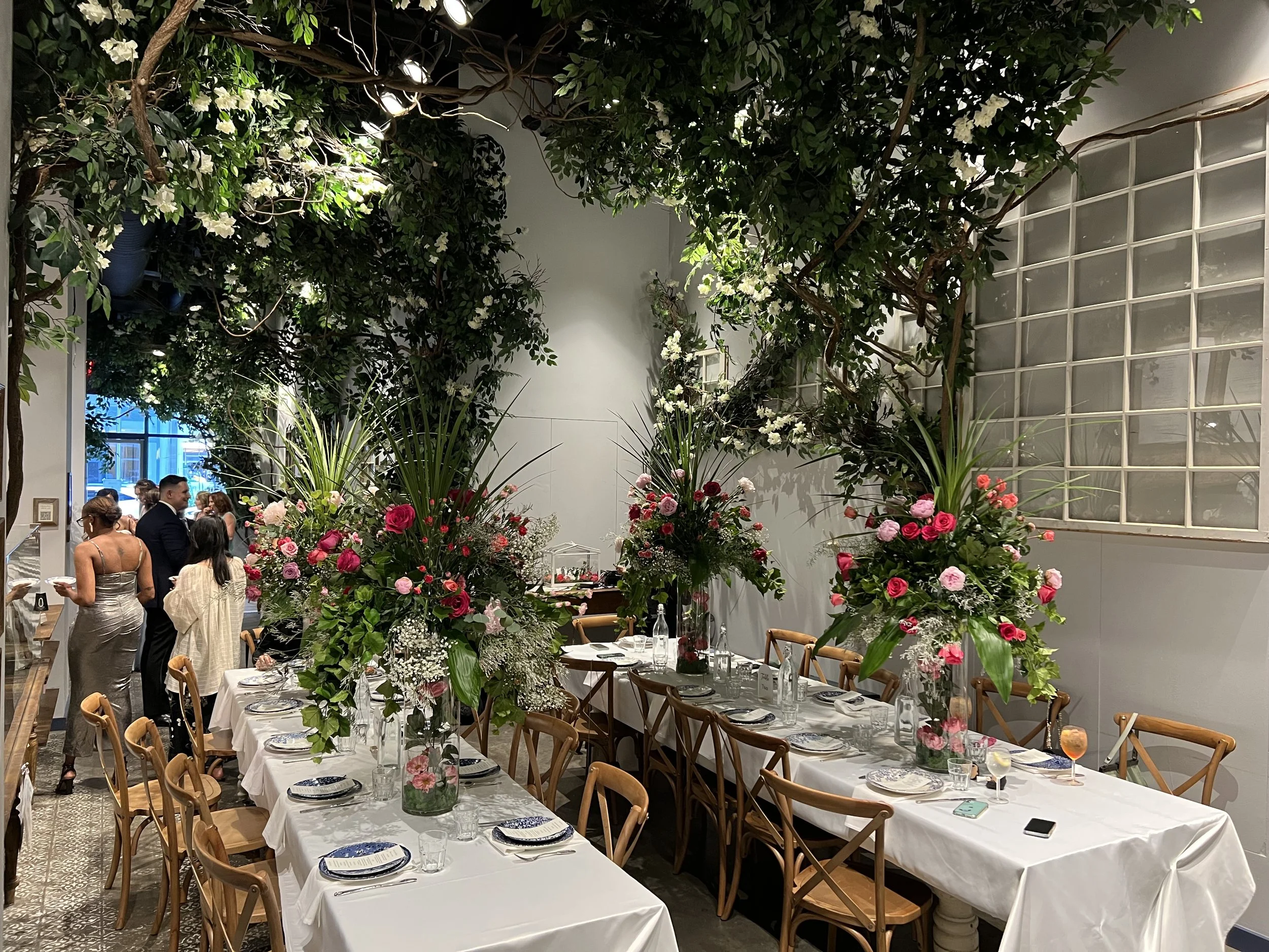 Dinner table setup with floral centerpieces, surrounded by wooden chairs, in a room decorated with greenery, flowers, and white walls, with people standing in the background.