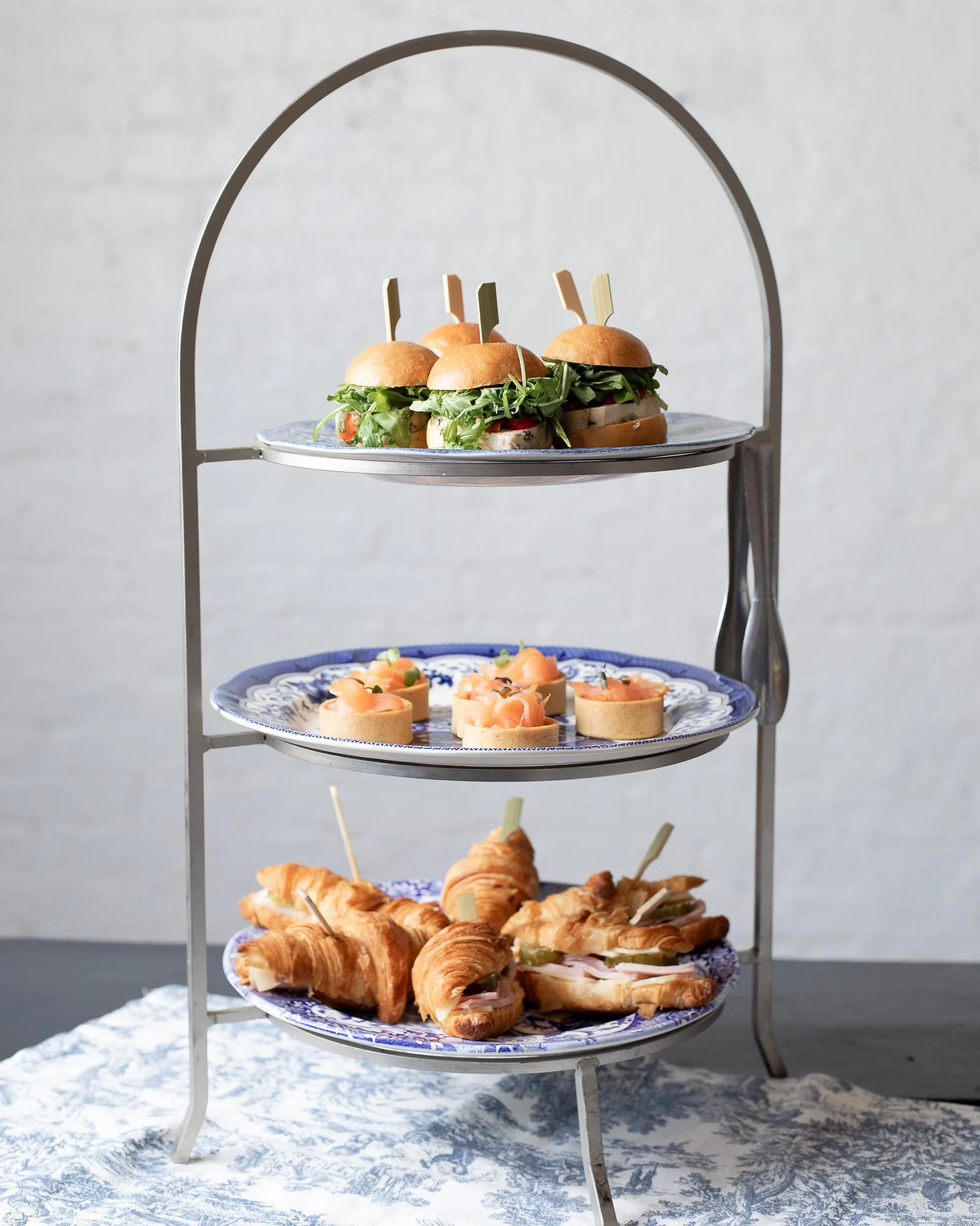 A tiered serving stand with three layers of appetizers: the top layer has small sandwiches with greens and toothpicks, the middle layer has small tartlets with smoked salmon, and the bottom layer has croissant sandwiches with meats and cheese.