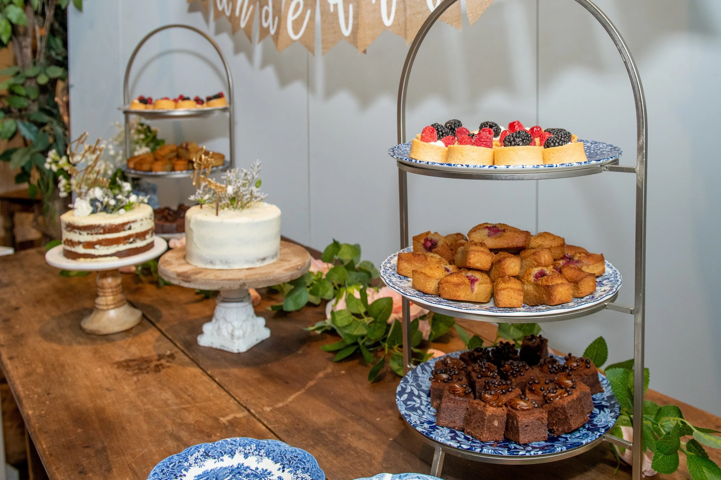 A dessert table with a variety of cakes, cookies, and pastries, along with a tiered stand holding fruit-topped desserts and cakes, and decorative greenery and a
