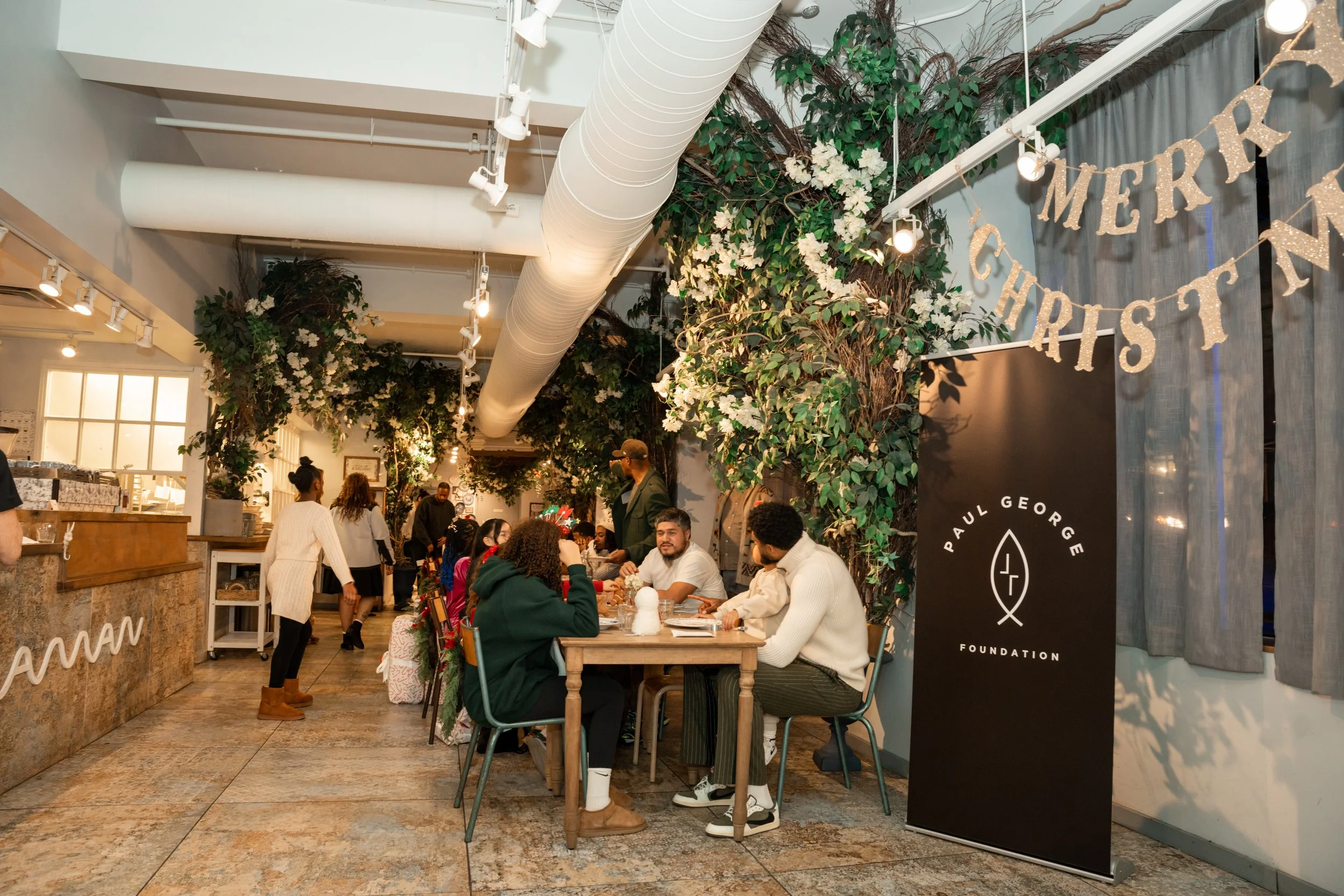 People sitting at a table inside a restaurant decorated with green plants and white flowers, during a Christmas celebration. There is a 'Merry Christmas' banner hanging and a sign with the Paul George Foundation logo.