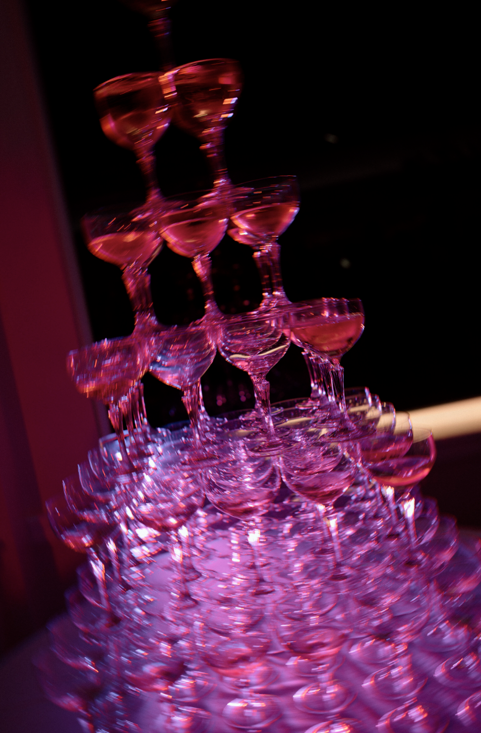 A pyramid of stacked pink cocktail glasses illuminated with pink lighting, set against a dark background.