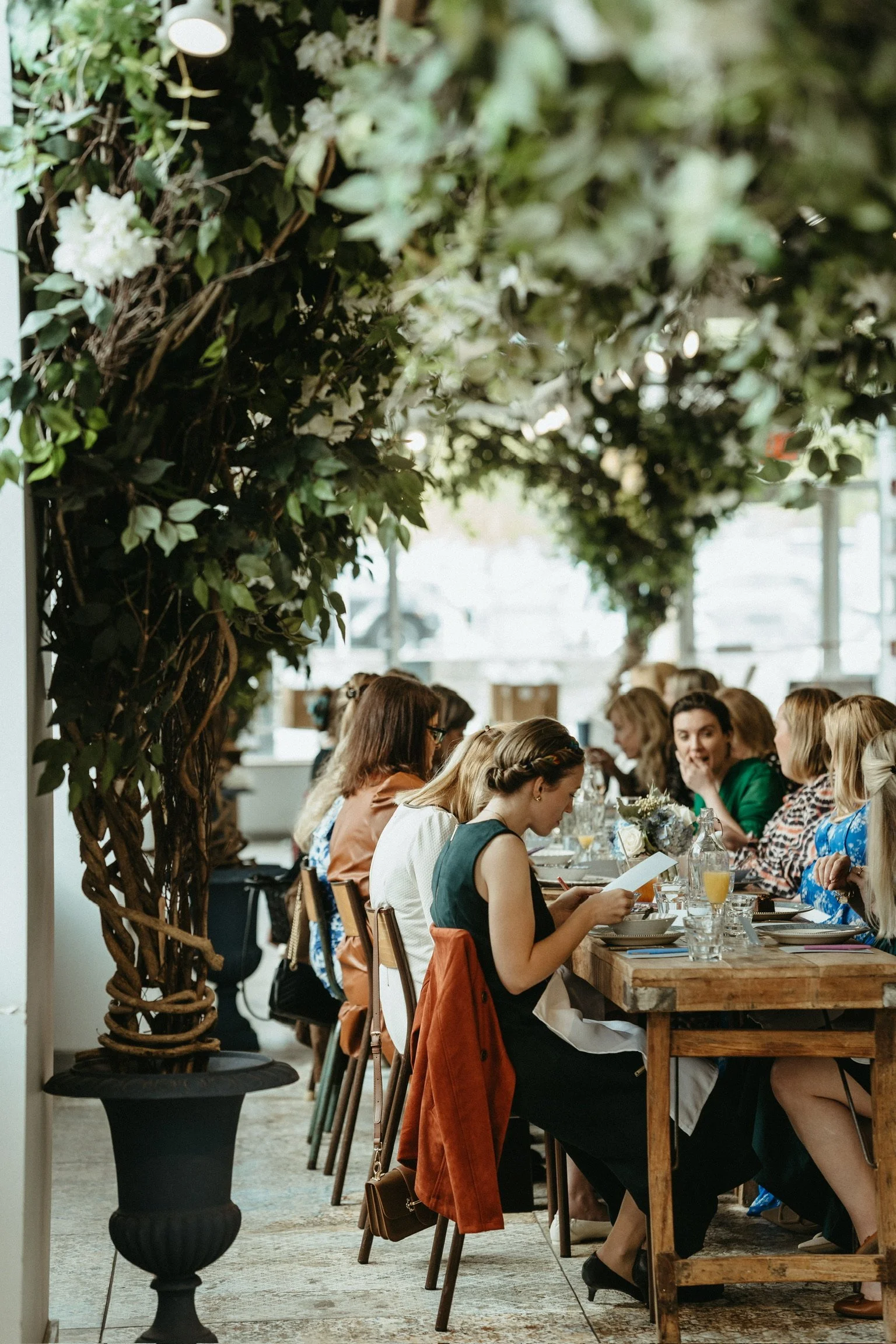 Women seated at a long dining table in a lush green indoor setting, engaged in conversation and enjoying drinks.