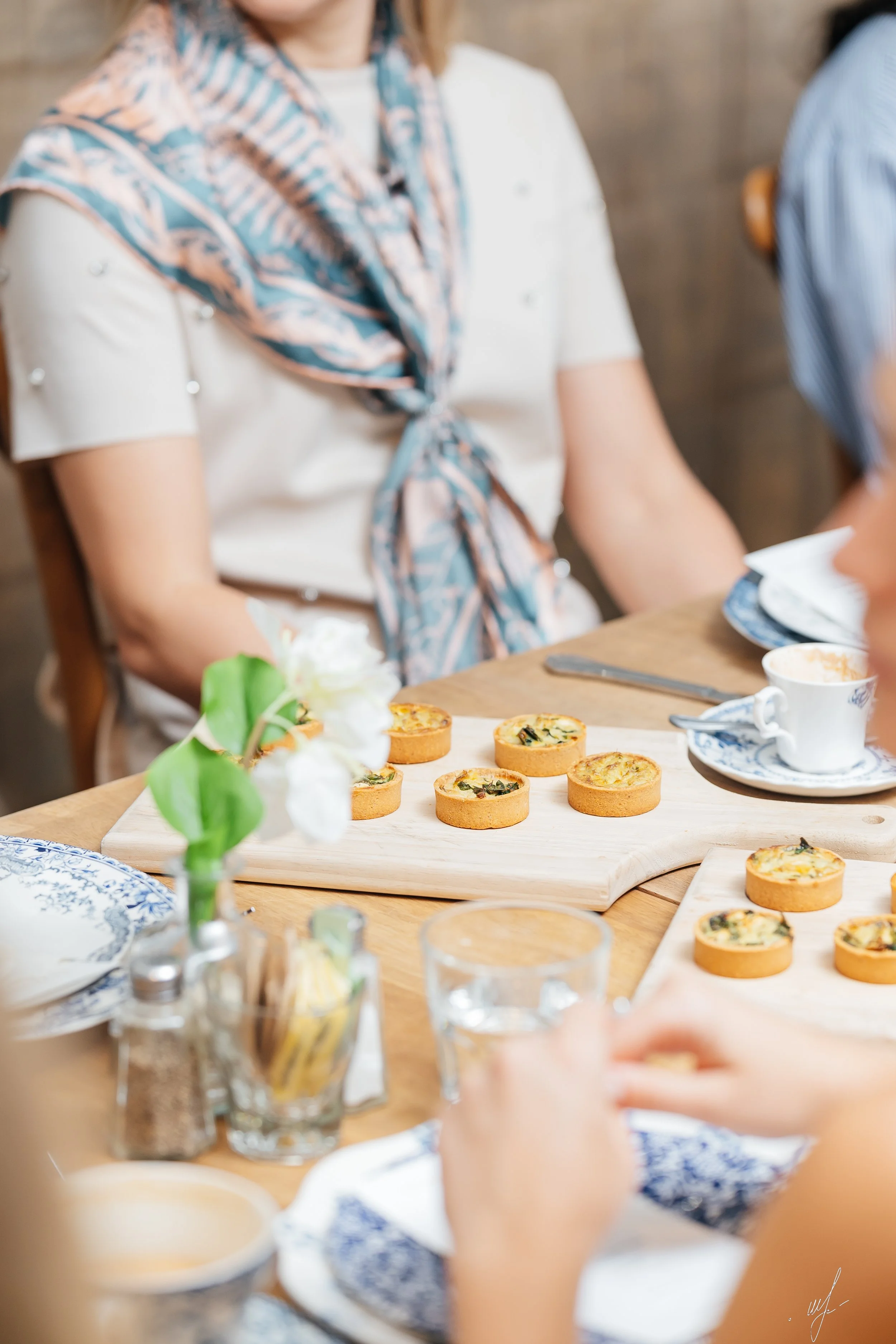 Close-up of a table with assorted tarts, cups, and glasses, with women sitting around, one in the background wearing a white top and a patterned scarf.