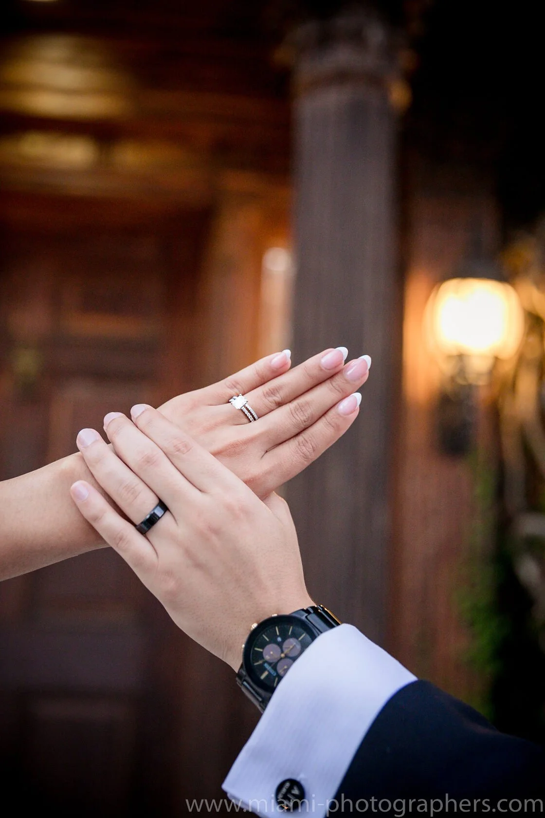 Close-up of a couple's hands showing their wedding rings, with a background of warm lighting and wooden pillars.