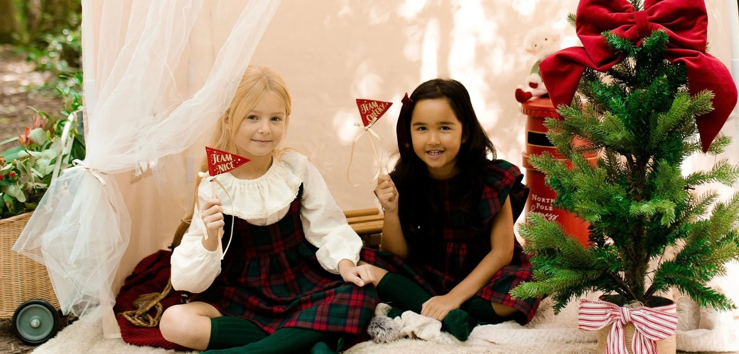 Two young girls sitting inside near a decorated Christmas tree, holding small signs that say 'Team Nice' and 'Team Naughty', with holiday decorations around them.