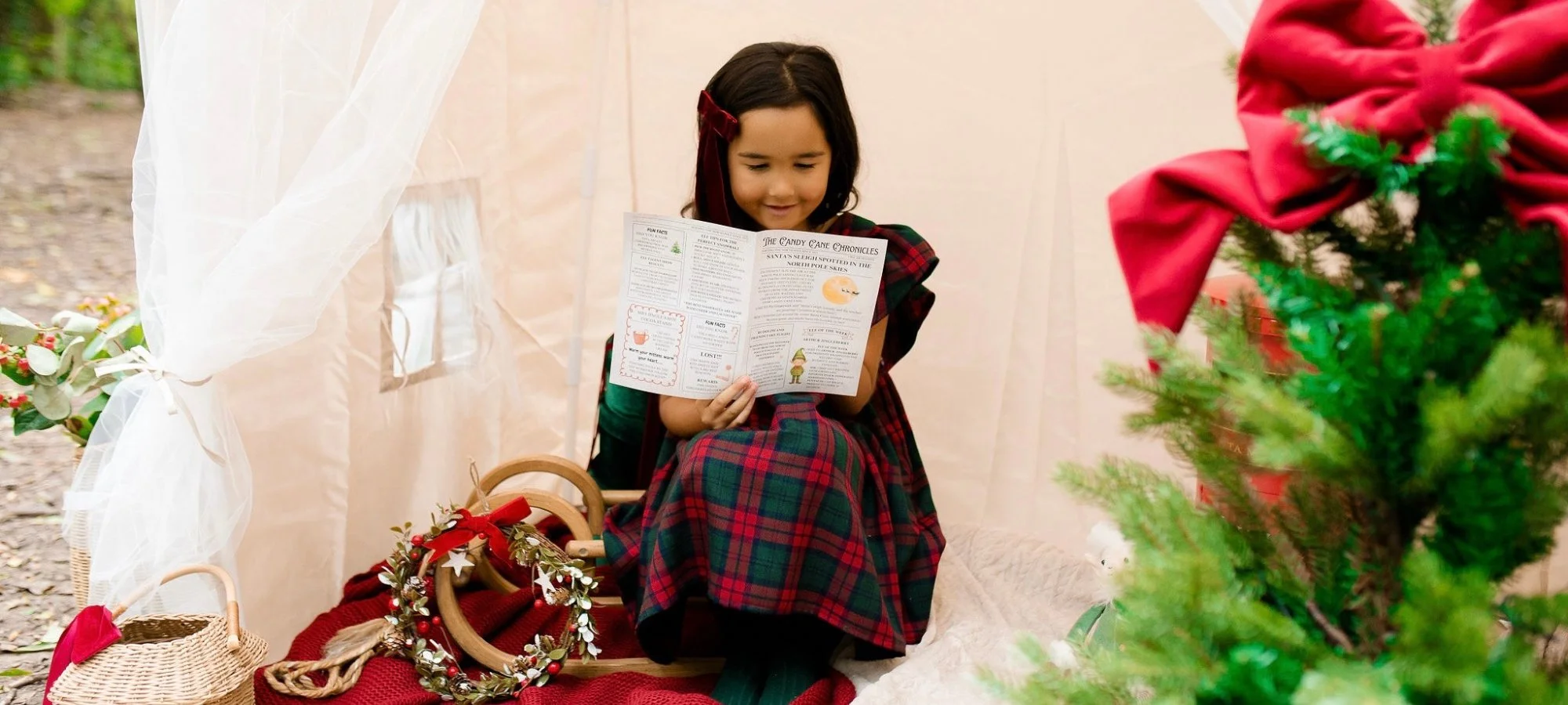 A young girl sitting inside a white tent decorated for Christmas, reading a holiday-themed flyer, with a Christmas tree and festive decorations nearby.