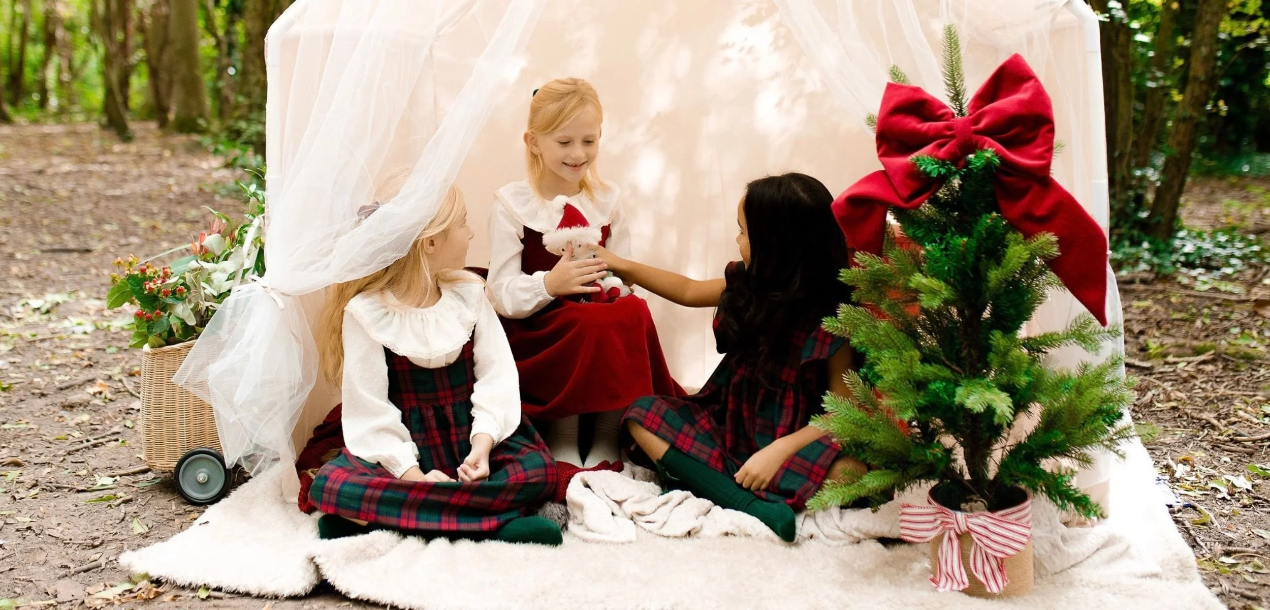 Four children dressed in Christmas outfits sitting outdoors on a blanket, engaged with a small girl wearing a Santa hat, amidst holiday decorations including a Christmas tree with a red bow, and a decorated basket of poinsettias.