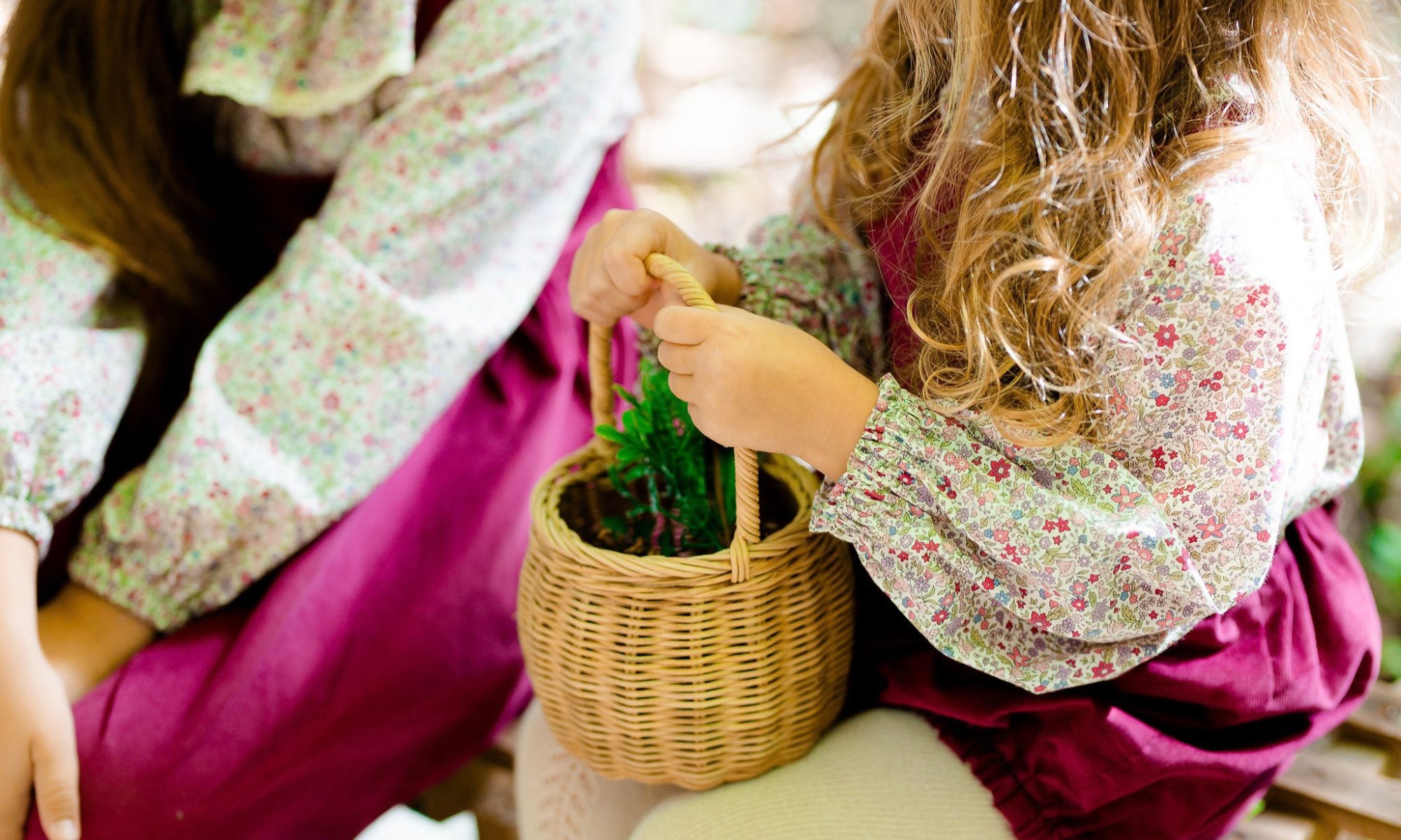 Children holding a small wicker basket with green artificial plants, wearing floral patterned clothing, sitting on a pink blanket.