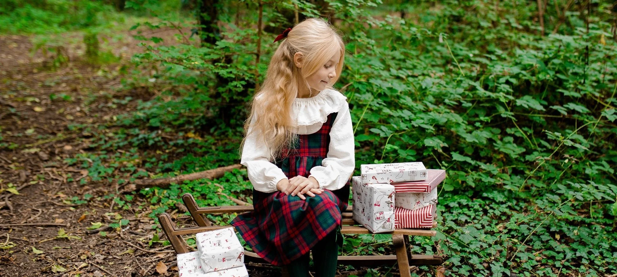 A young girl with long curly blonde hair sitting on a wooden bench in a forest, surrounded by green foliage, with wrapped Christmas presents in front of her.