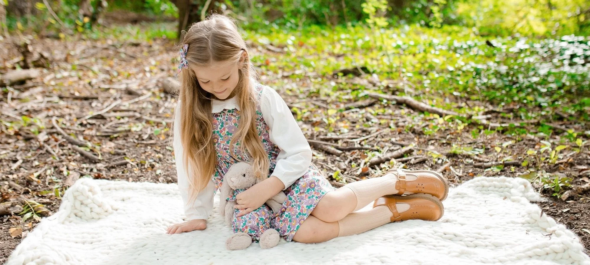 A young girl wearing a floral dress, white cardigan, and tan shoes, sitting on a white blanket in a wooded area. She is holding a stuffed bunny toy and looking down at it.
