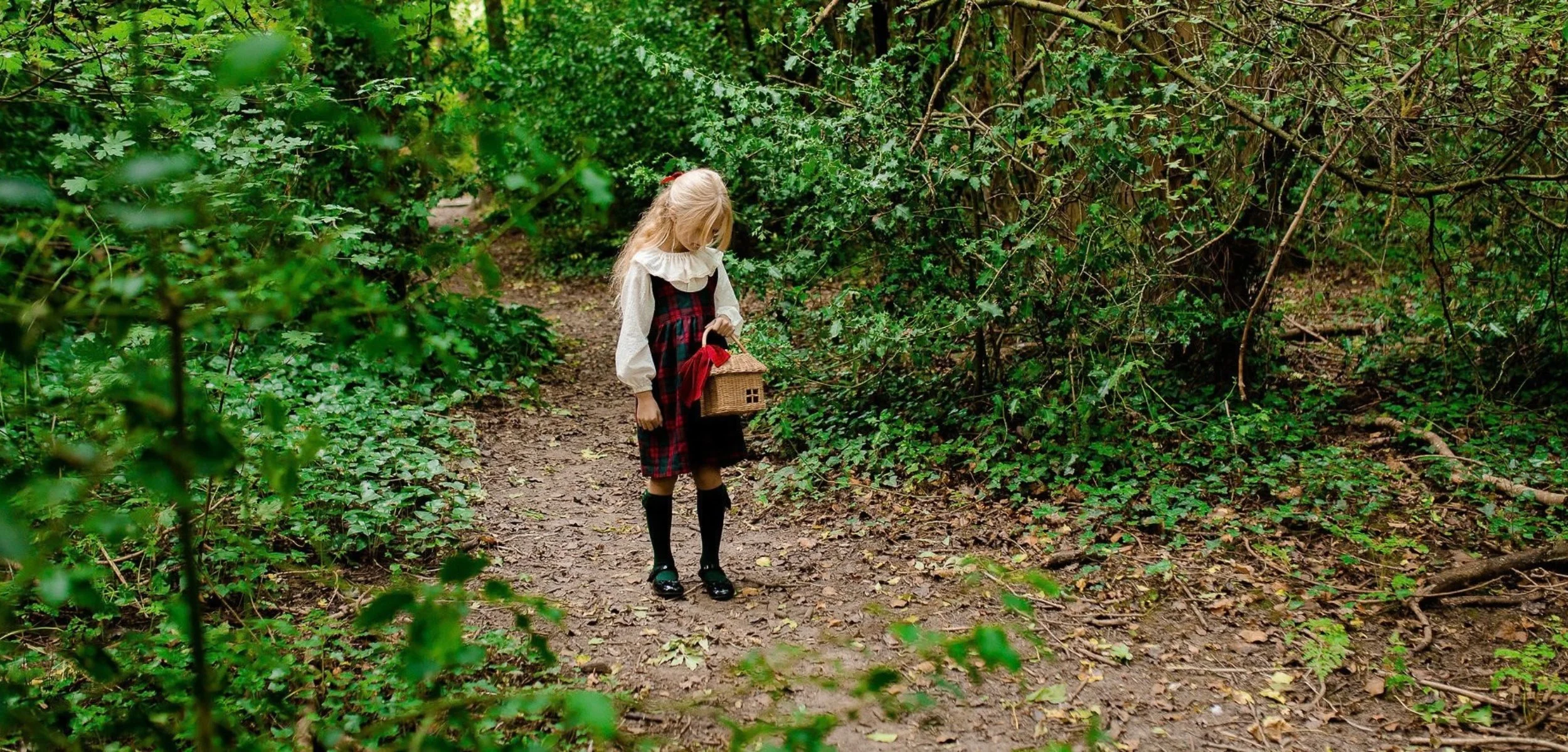 A young girl with long blonde hair dressed in vintage-style clothing, holding a small wicker house-shaped basket, standing on a dirt path in a lush green forest.