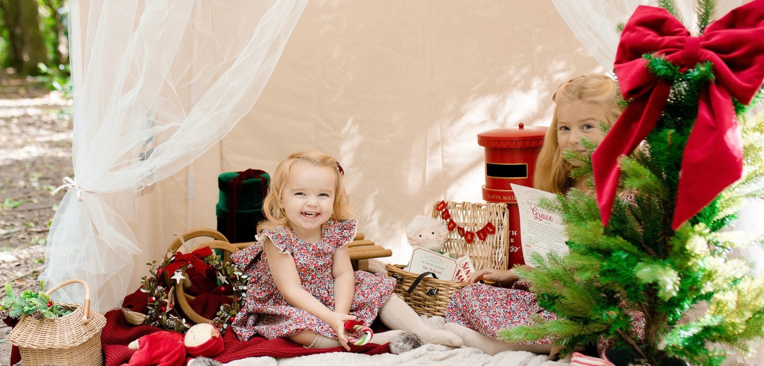 Two young girls sitting inside a decorated Christmas-themed setting surrounded by holiday decorations including a small Christmas tree, gift baskets, and a red mailbox. One girl is smiling at the camera, while the other is partially visible behind a 