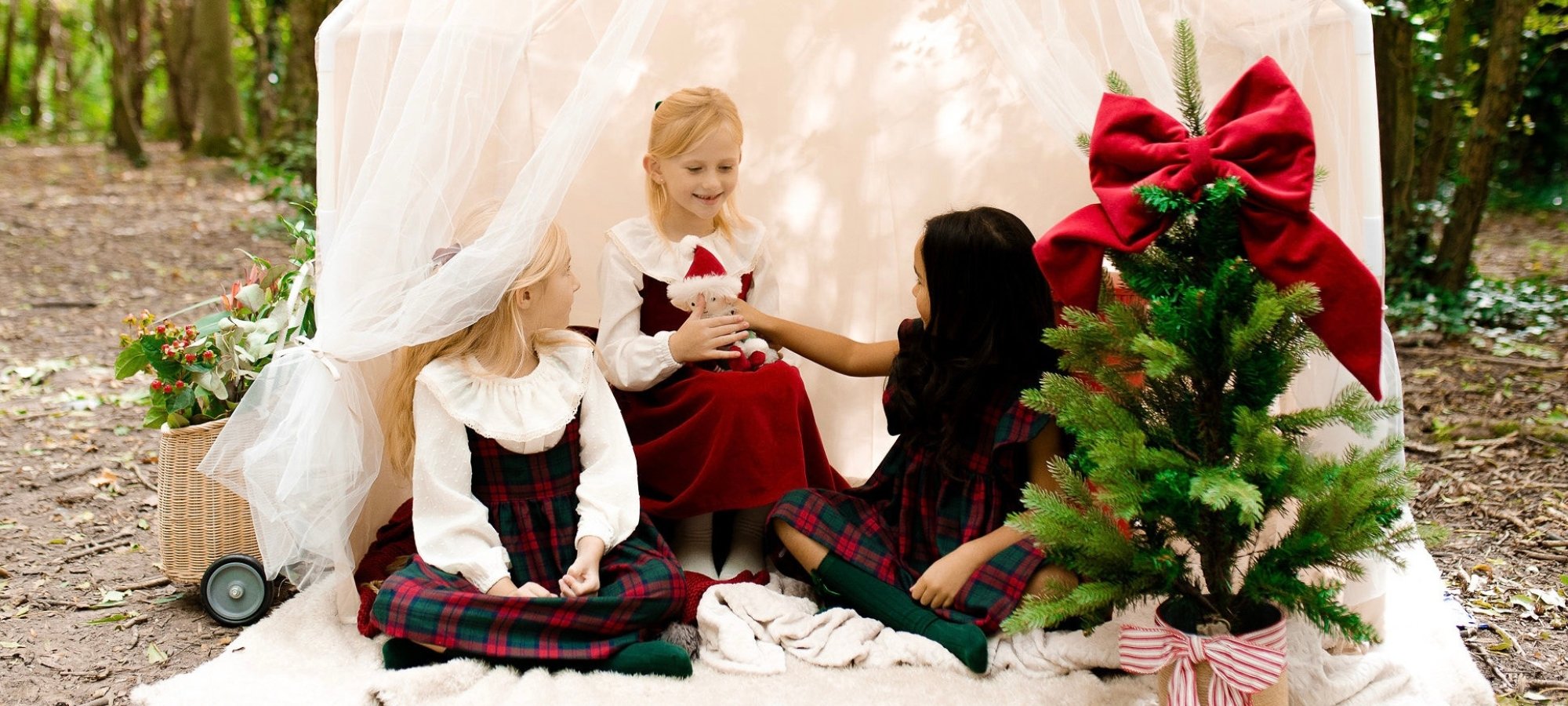 Four young girls in festive red and green Christmas dresses participating in a nativity scene outdoors, with one girl dressed as Mary, holding a stuffed sheep, and the others gathered around, near a decorated small Christmas tree and a basket of flowers.