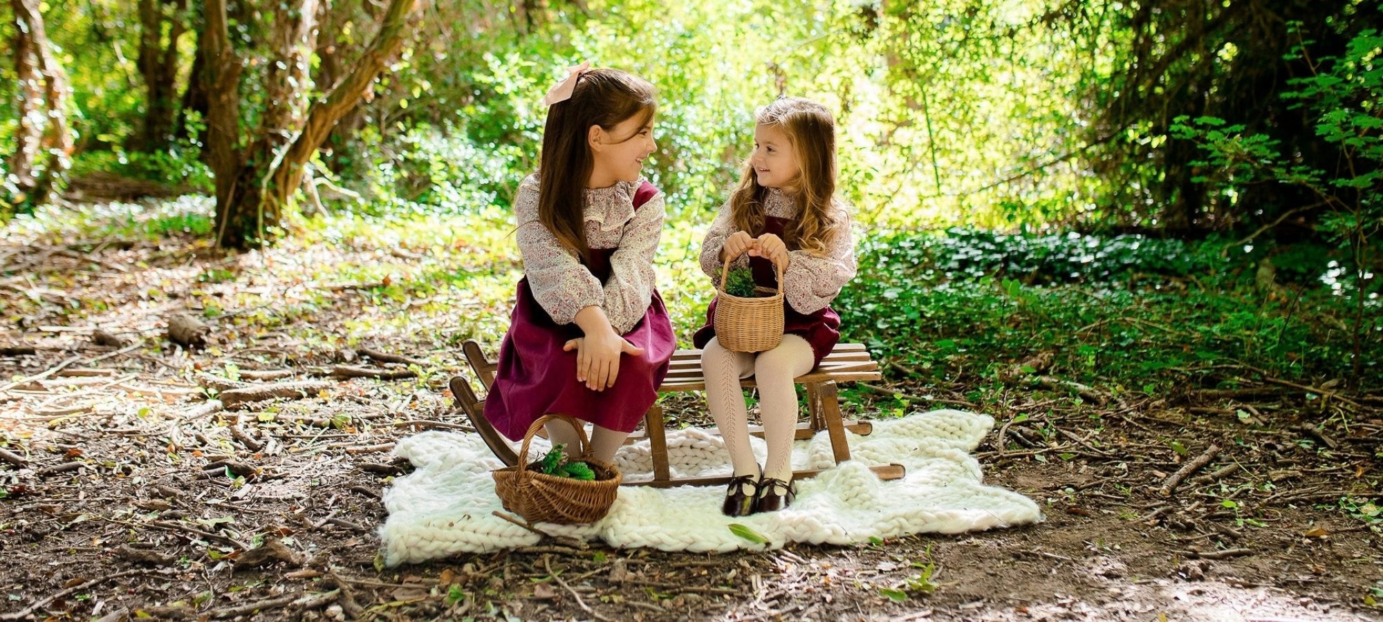 Two girls sitting on a wooden bench in a forest, sharing a moment with one holding a basket. They are dressed in vintage-style clothing, surrounded by greenery with sunlight filtering through the trees.