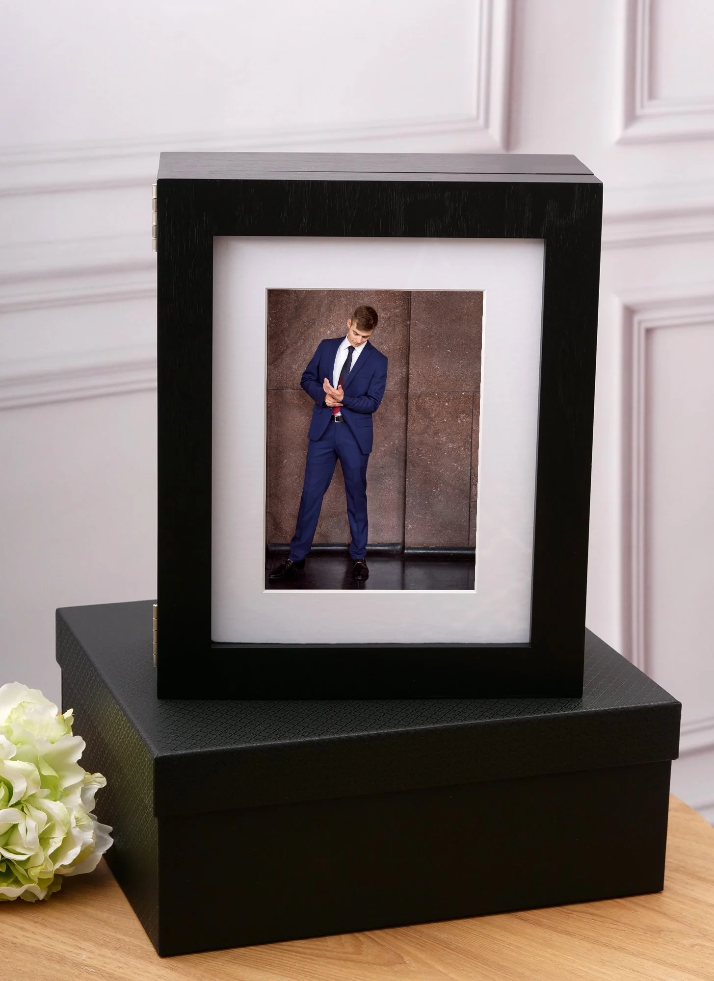 A framed photograph of a young man in a blue suit standing against a brown wall, inside a black picture frame resting on a black box, with white flowers nearby.