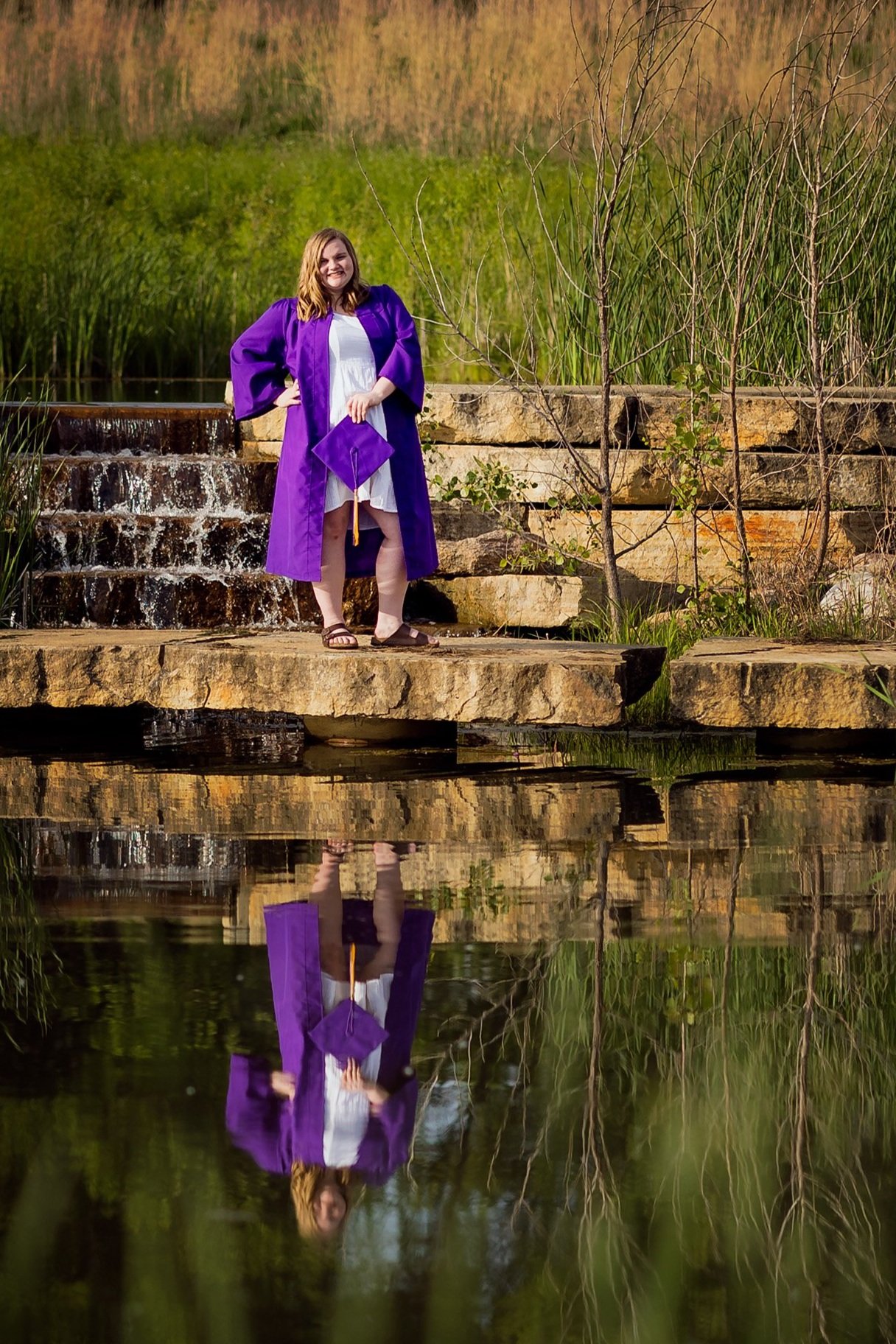 A young woman in a white dress and purple graduation gown holding a graduation cap and standing on rocks near a small waterfall and pond, with her reflection visible in the water.