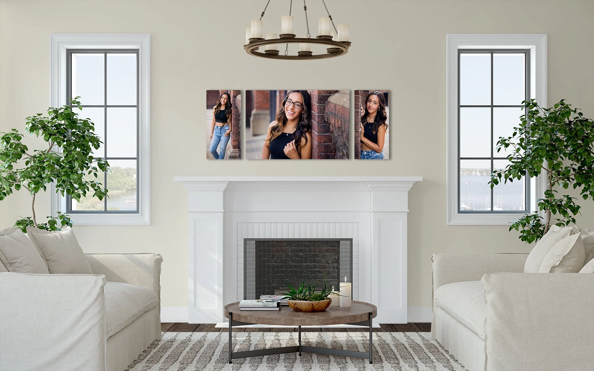Living room with beige sofas, a round wooden coffee table with a plant and books, two large windows showing a water view, and a white fireplace with a triptych of photographs of a smiling young woman on the wall above.