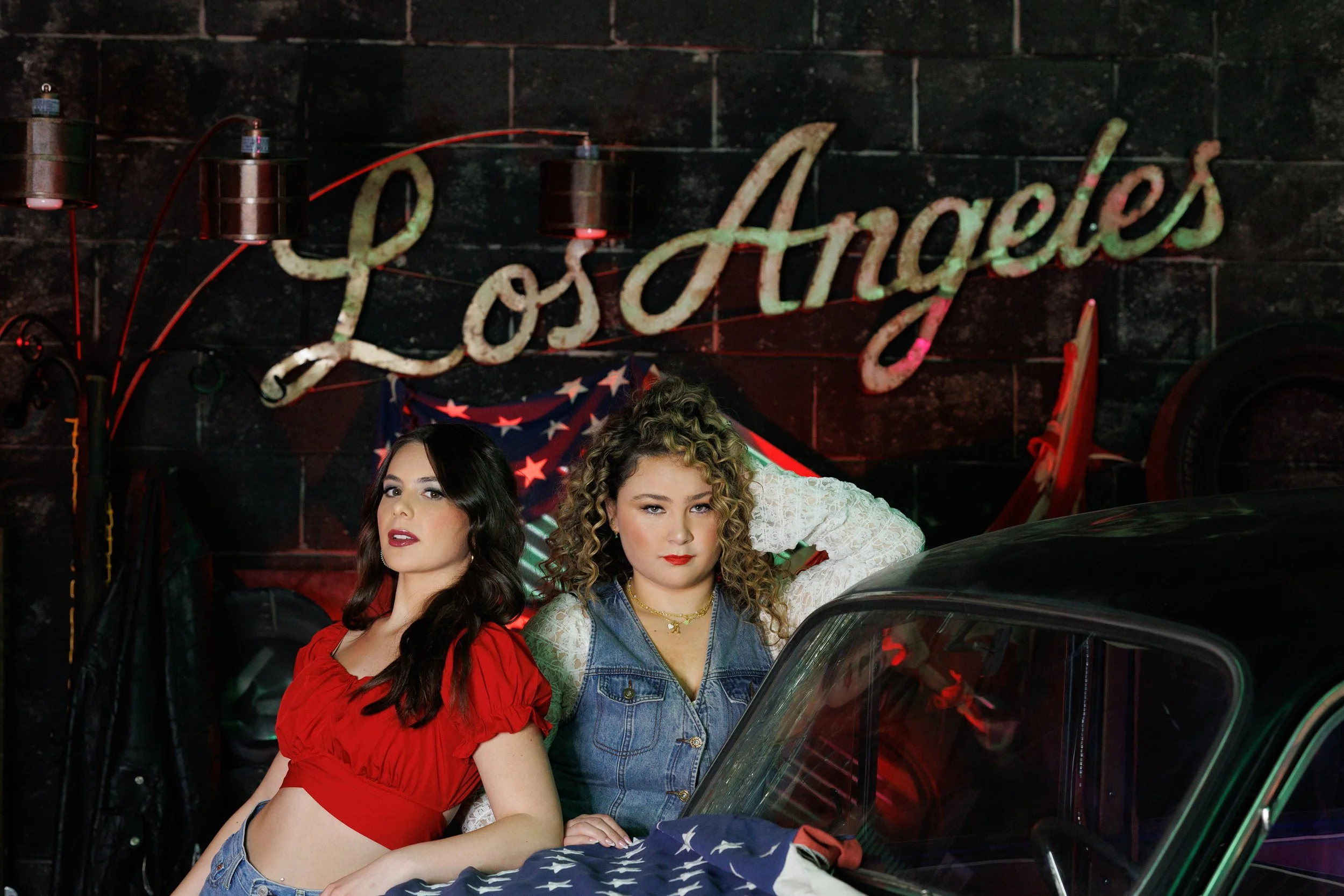 Two high school senior girls lean on a Volkswagon Bug dressed in red, white, and blue with a lit Los Angeles sign behind them.