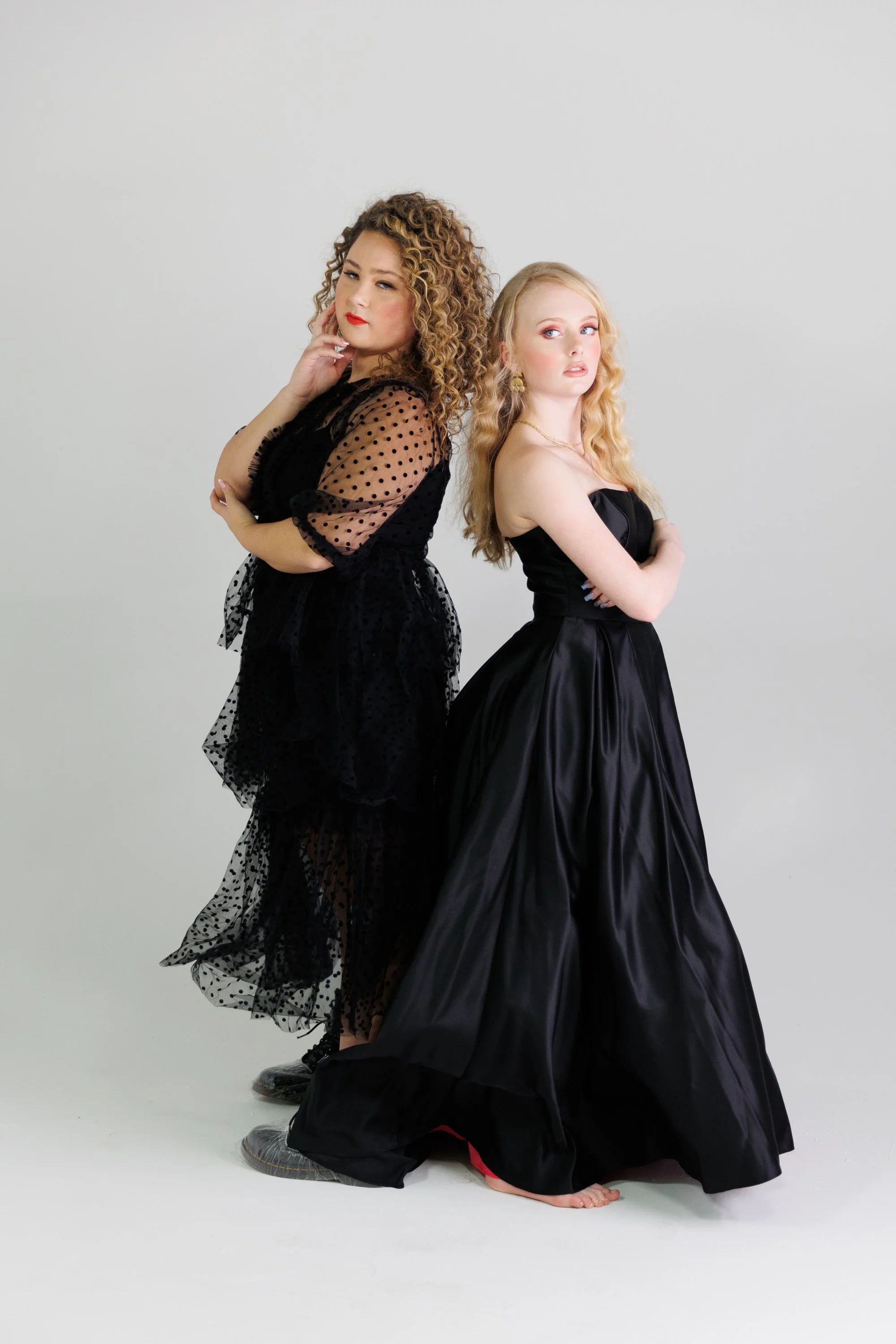 Two high school seniors dressed in formal black dresses pose back to back against a white background.