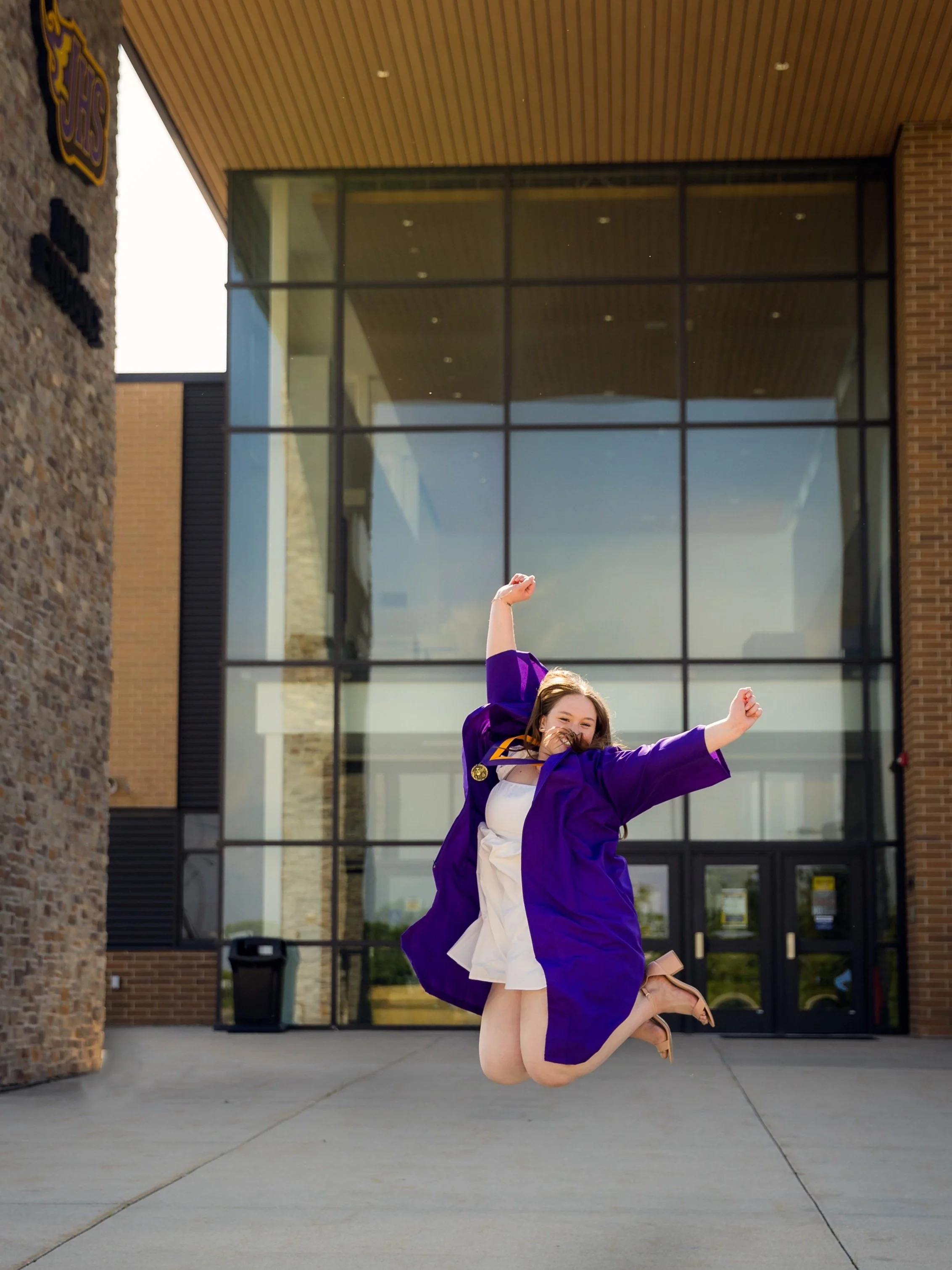 A young woman in a graduation gown jumping in the air outside a modern building with large glass windows.