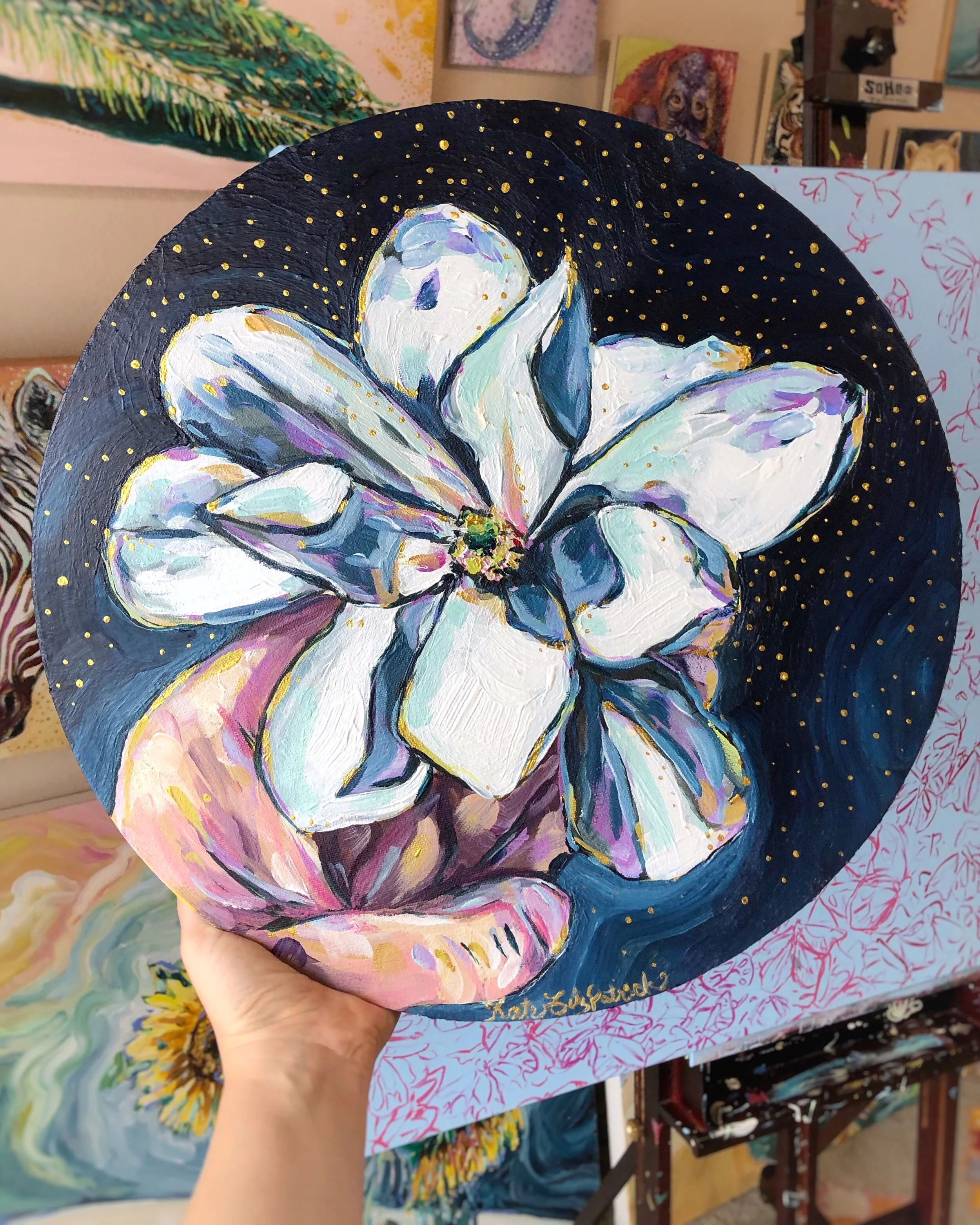 A round canvas is held in a white hand. On the canvas is a painting of a magnolia flower, also held in a white hand, against a deep blue background.