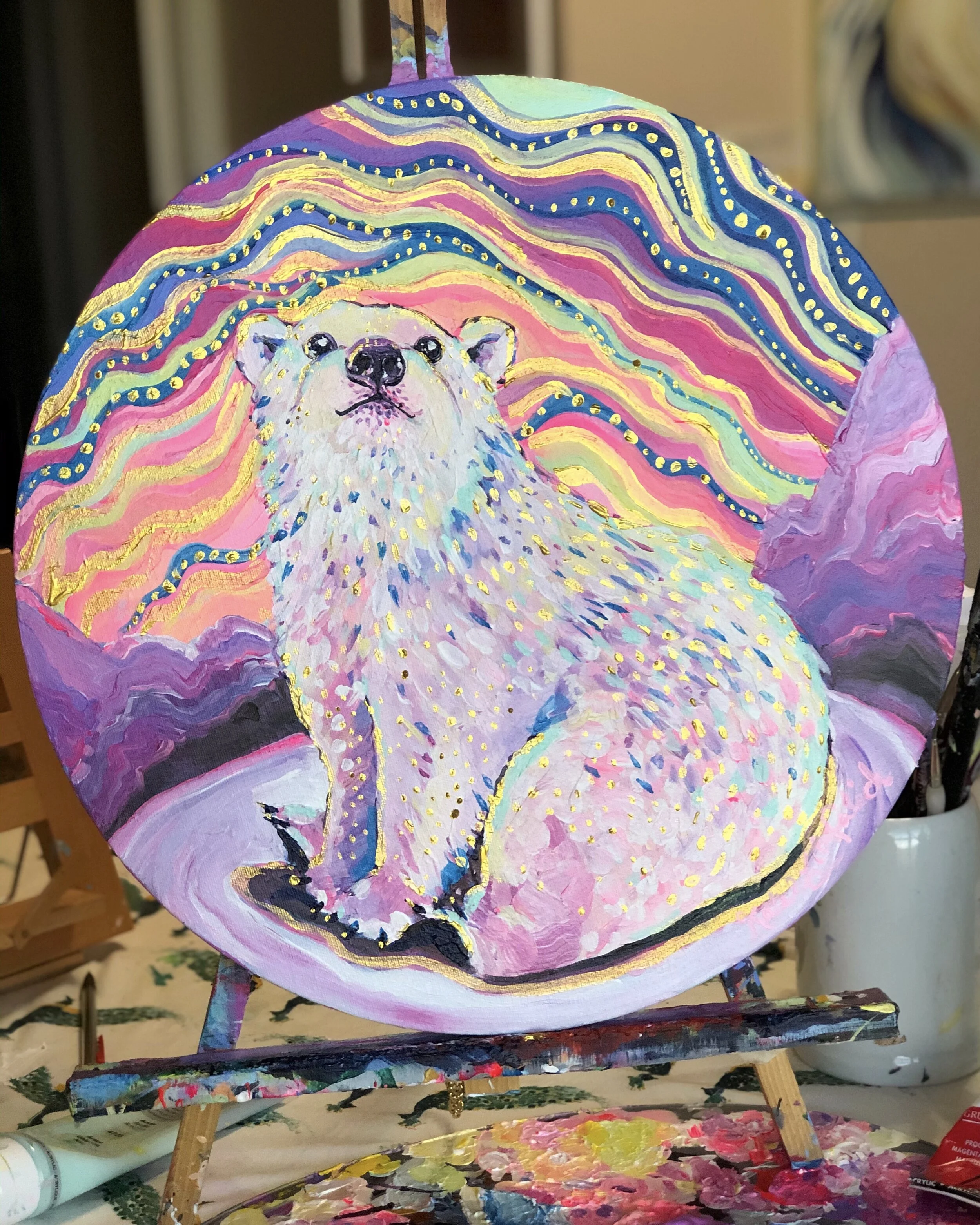 A painting of a polar bear cub against a colorful sky, like the aurora borealis. The canvas is round and sitting on a small easel.