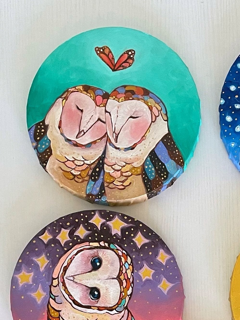 a round painting of two barn owls leaning in to each other with a butterfly in the shape of a heart above them