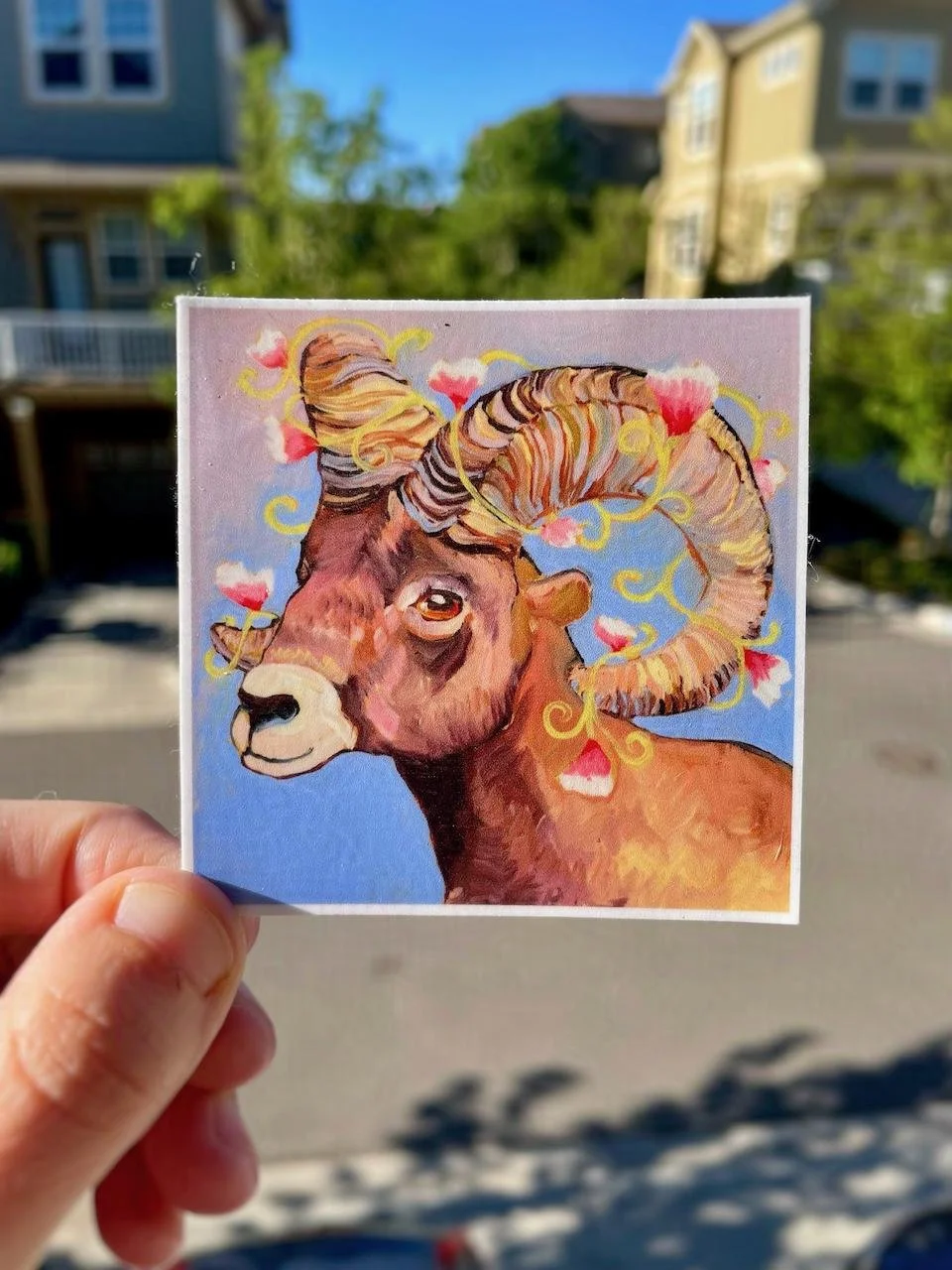a square sticker of a bighorn sheep painting. there are flowers and vines around the sheep's horns