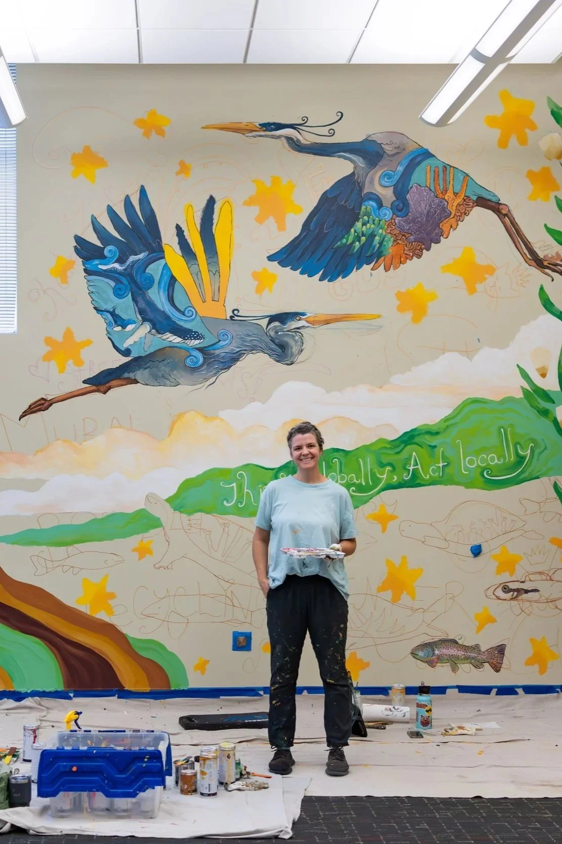 Artist standing in front of a colorful mural of flying herons, yellow maple leaves, and aquatic animals with a green banner that says "They Are Really Act Locally." The artist is holding a palette and is surrounded by painting supplies on the floor.