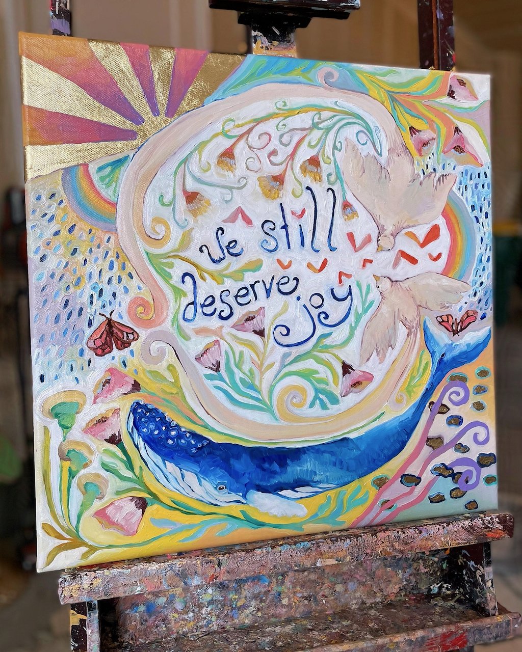 a colorful painting with a bird, flowers, a whale, and rainbows. the words 'we still deserve joy' are painted in the center.