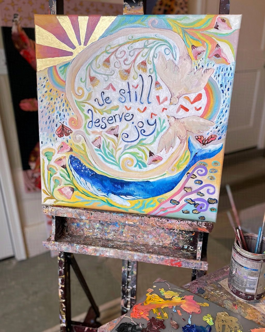 a colorful painting with a bird, flowers, a whale, and rainbows. the words 'we still deserve joy' are painted in the center.