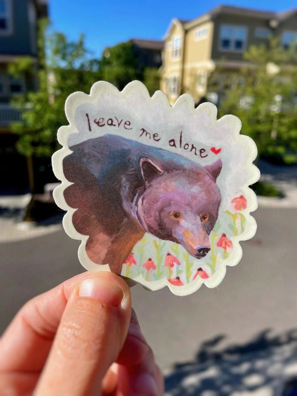 a sticker of a bear among flowers with the words 'leave me alone' and a hear beside it. the sticker is oval shaped with scalloped edges.