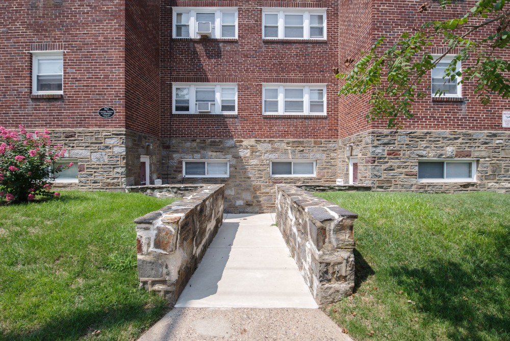 Concrete walkway with stone railings leading to the entrance of a brick apartment building with small grass lawn and flowering shrub on the left.