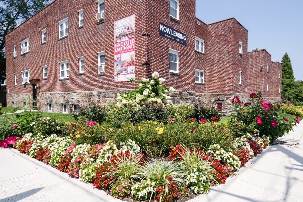 A brick apartment building with signs indicating it is for rent and lease, surrounded by a garden with colorful flowers.