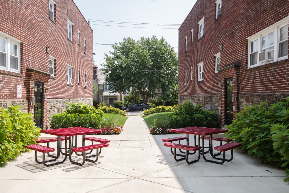 Outdoor courtyard between two red brick apartment buildings with red picnic tables, green bushes, flowers, and a large tree in the background.
