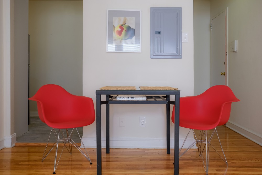 A small dining area with a black table, two red modern chairs, a framed colorful still life painting on the wall, and an electrical panel on the wall.