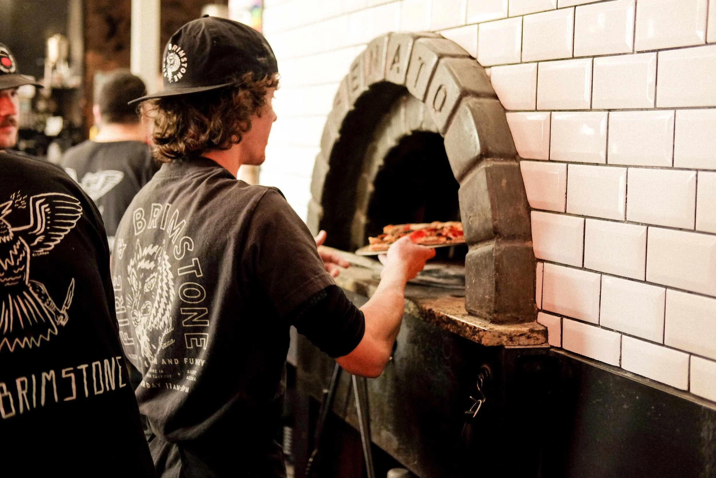 Chef at Fire and Brimstone placing pizza in wood fired oven wearing branded merch and hat designs