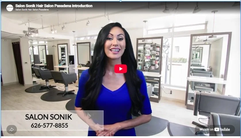 Woman with long black hair smiling inside Salon Sonik in Pasadena, with styling stations, mirrors, salon chairs and shelves of hair products.