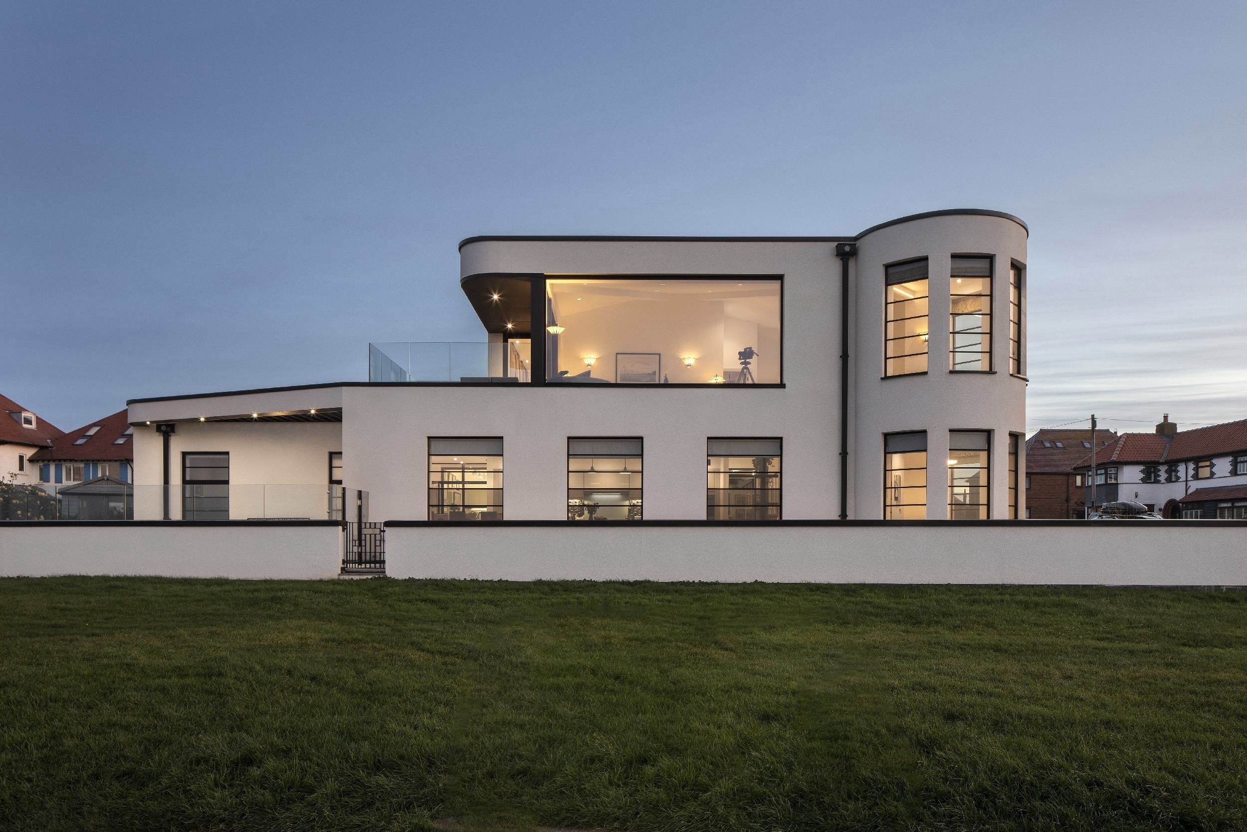White Art Deco house, dominated by a large picture window on the first floor. Modern turret with panoramic windows. Cloudless sunset sky behind. Property is framed by a low white wall.
