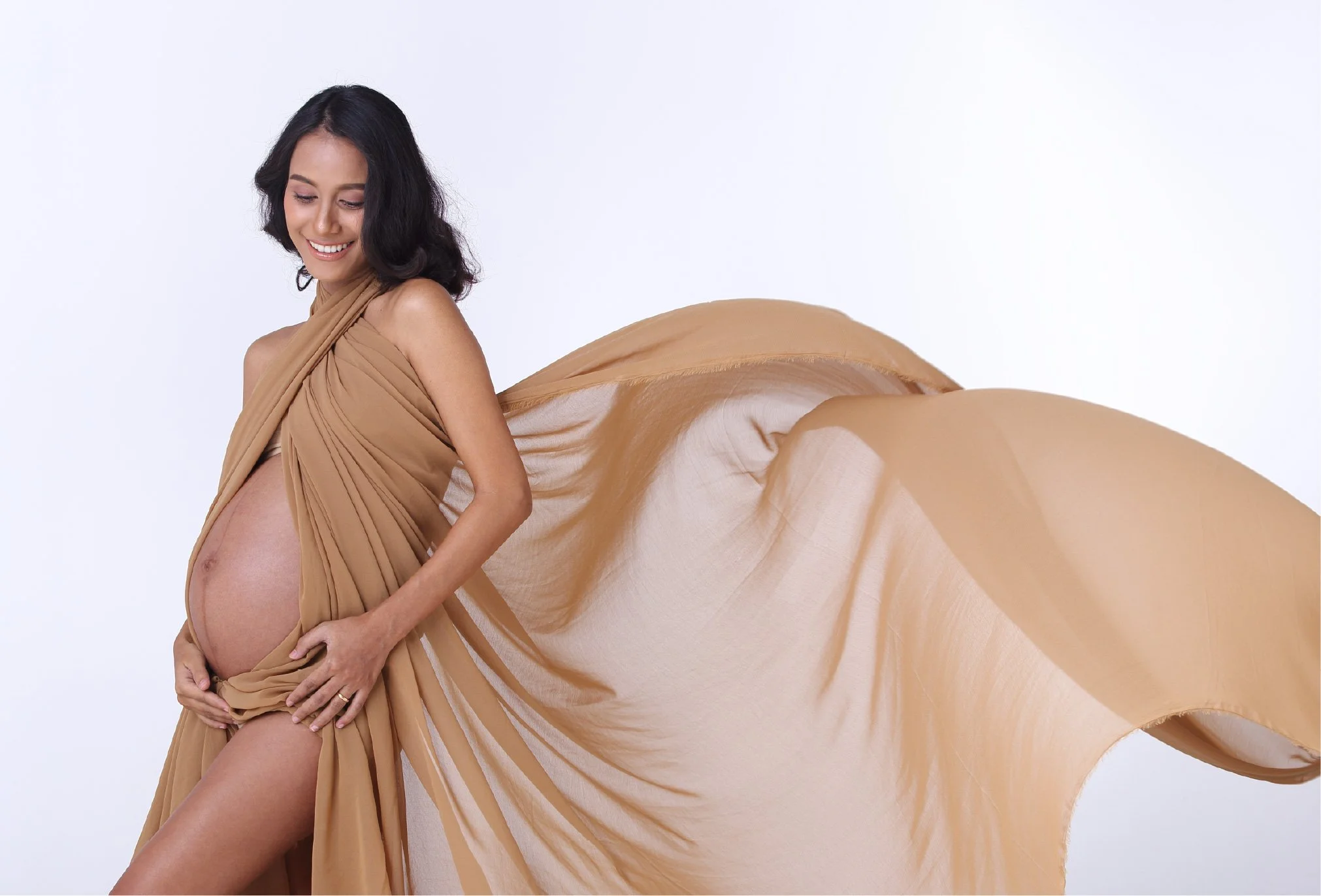 A pregnant woman with dark hair smiling while holding her belly, dressed in flowing beige fabric against a plain white background.