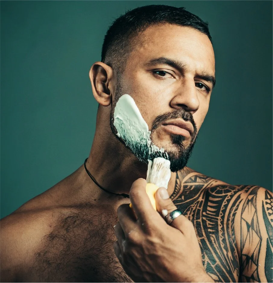 A shirtless man with tattoos on his arm and shoulder, wearing a black necklace, is holding a shaving brush and applying shaving cream to his face.
